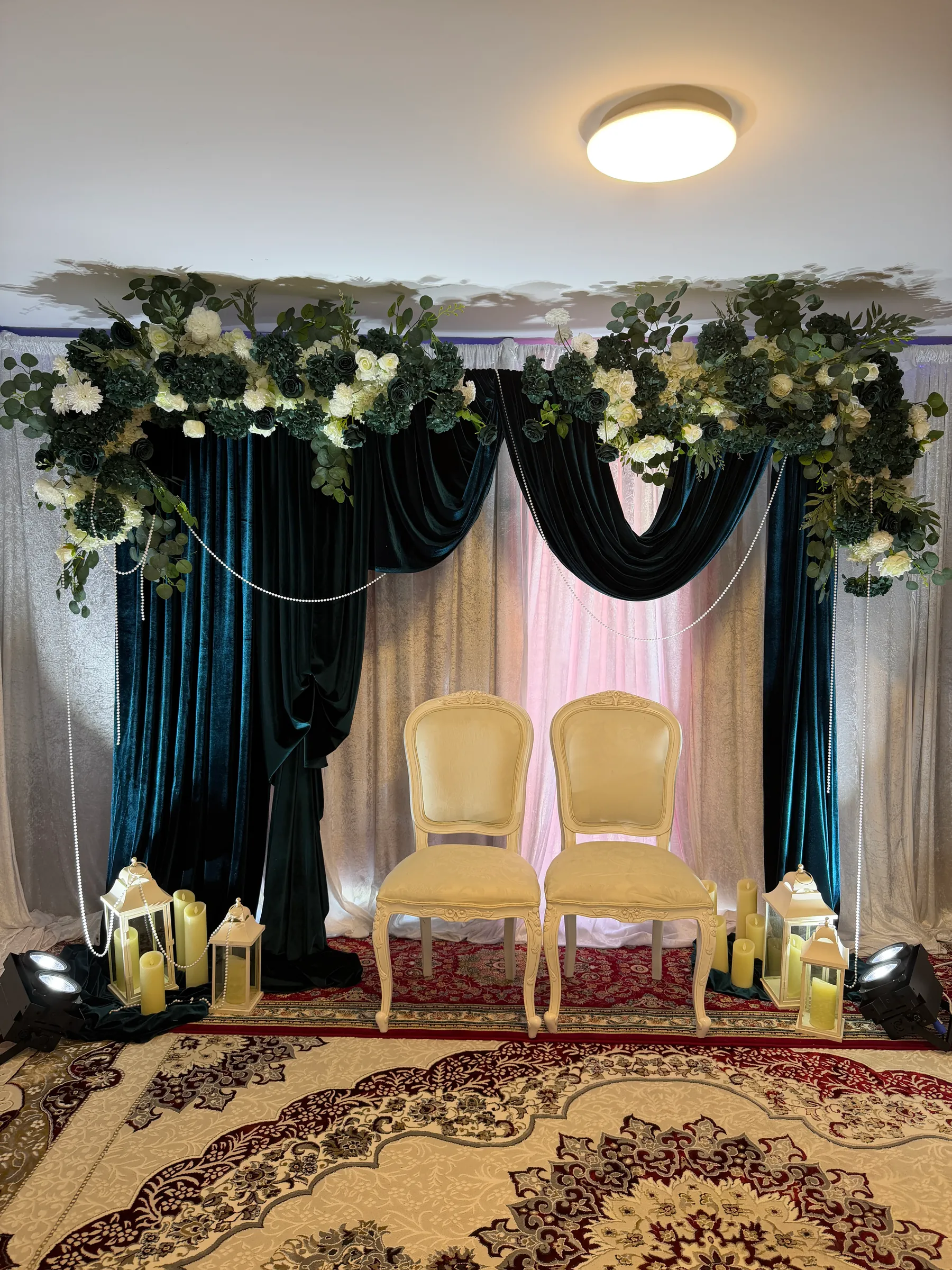 Stage styling by Decoration X: Two cream carved chairs on a Persian rug before teal velvet swags, white drapes and greenery and white floral clusters, candle lanterns at each side.
