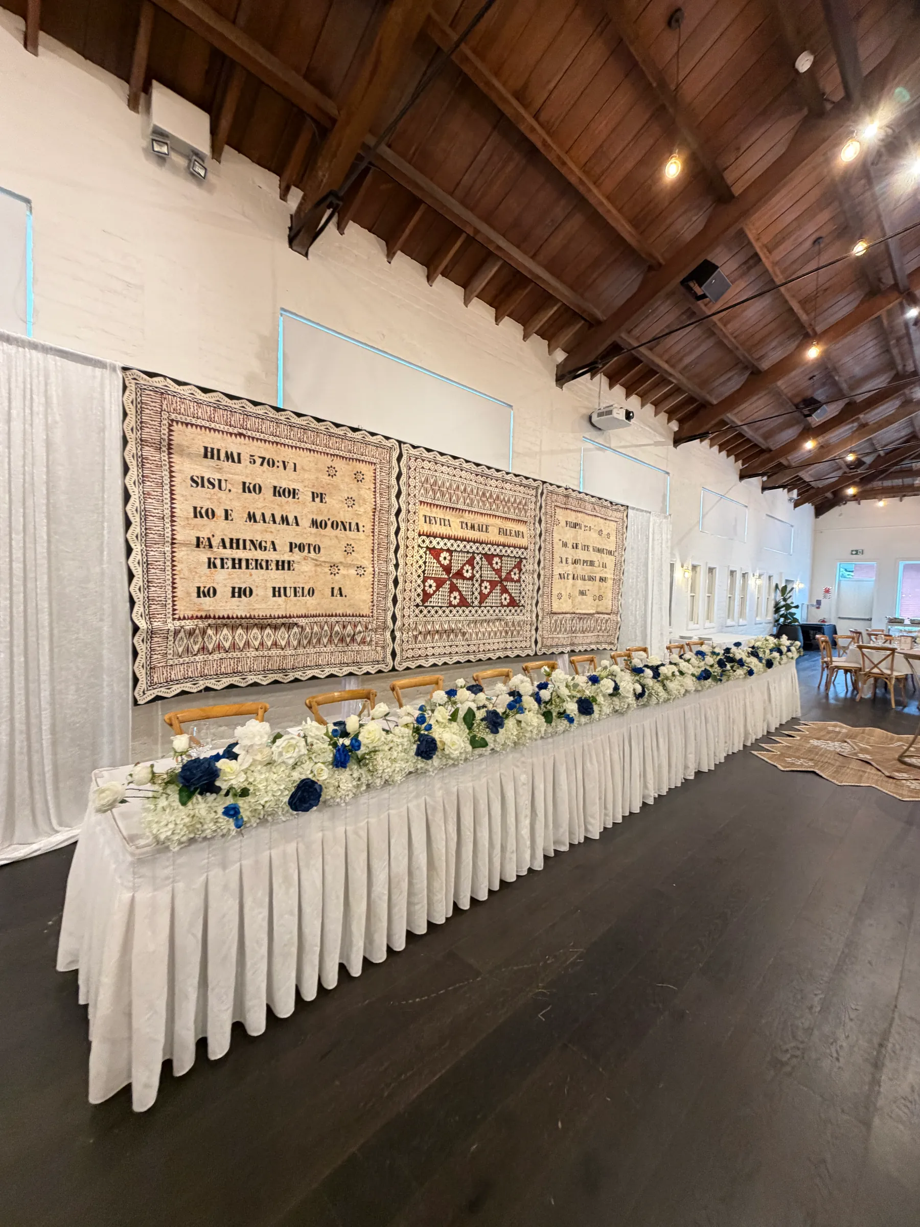 Cultural styling by Decoration X: Skirted head table with a white hydrangea and navy rose runner set before three hanging tapa cloths printed with hymn and scripture text in Tongan.