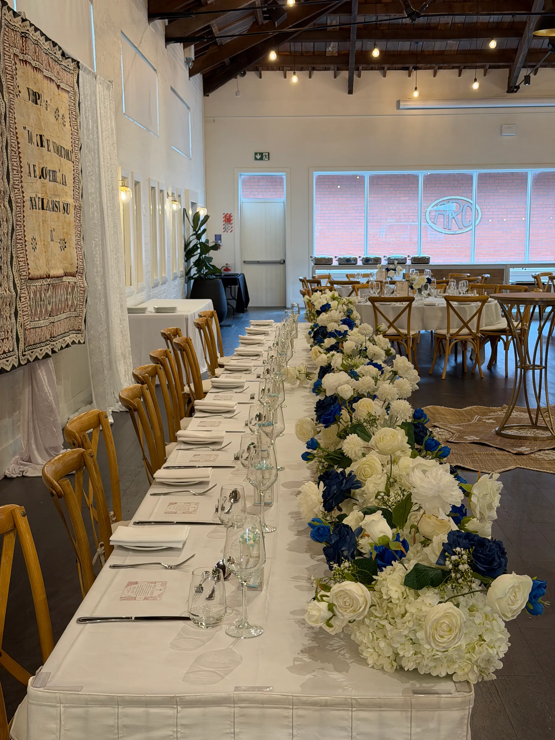 Tables styling by Decoration X: Long table runner of white roses, hydrangea and navy roses beside place settings, glassware and folded napkins, crossback chairs along one side.