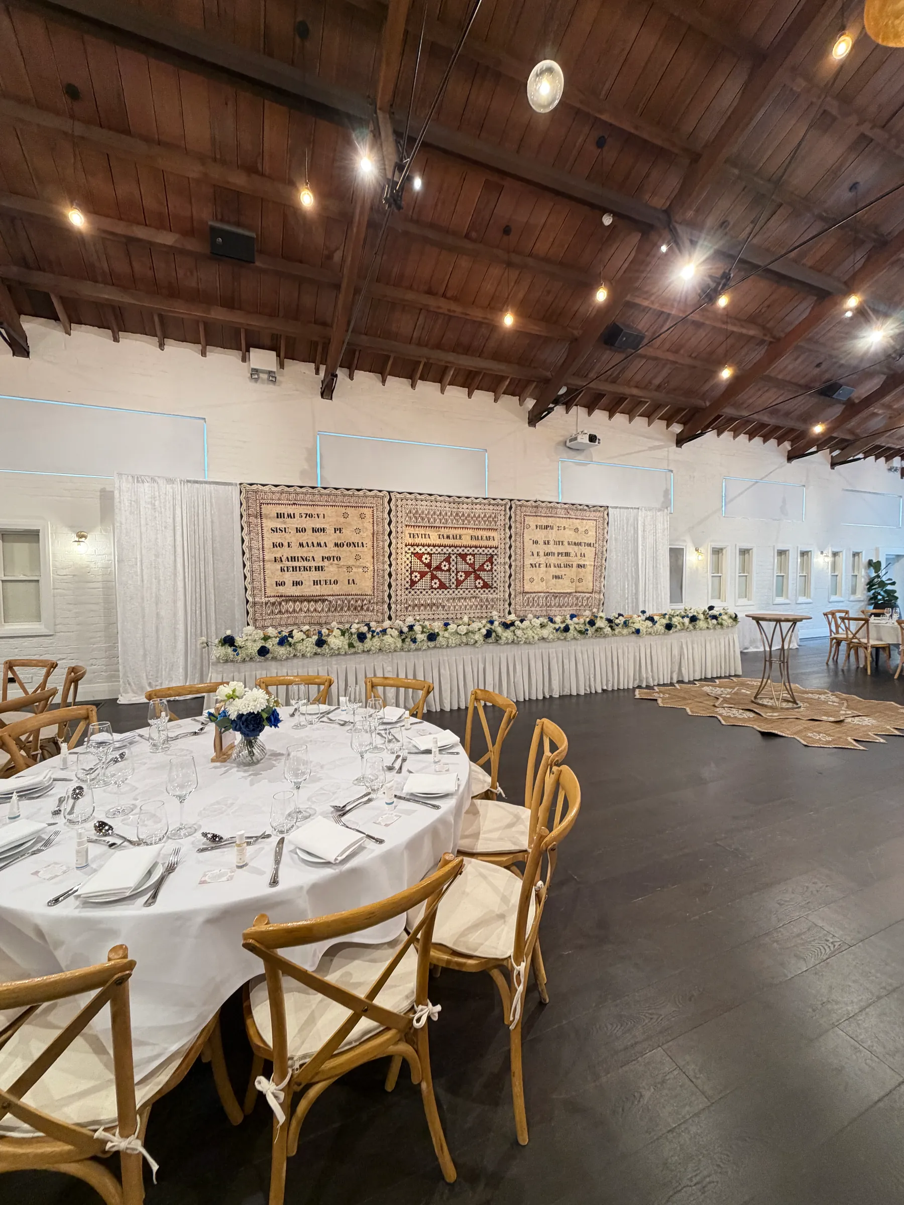 Reception styling by Decoration X: Round guest table with crossback chairs and a blue and white posy, a long skirted head table with blue and white florals before three hanging tapa cloths.