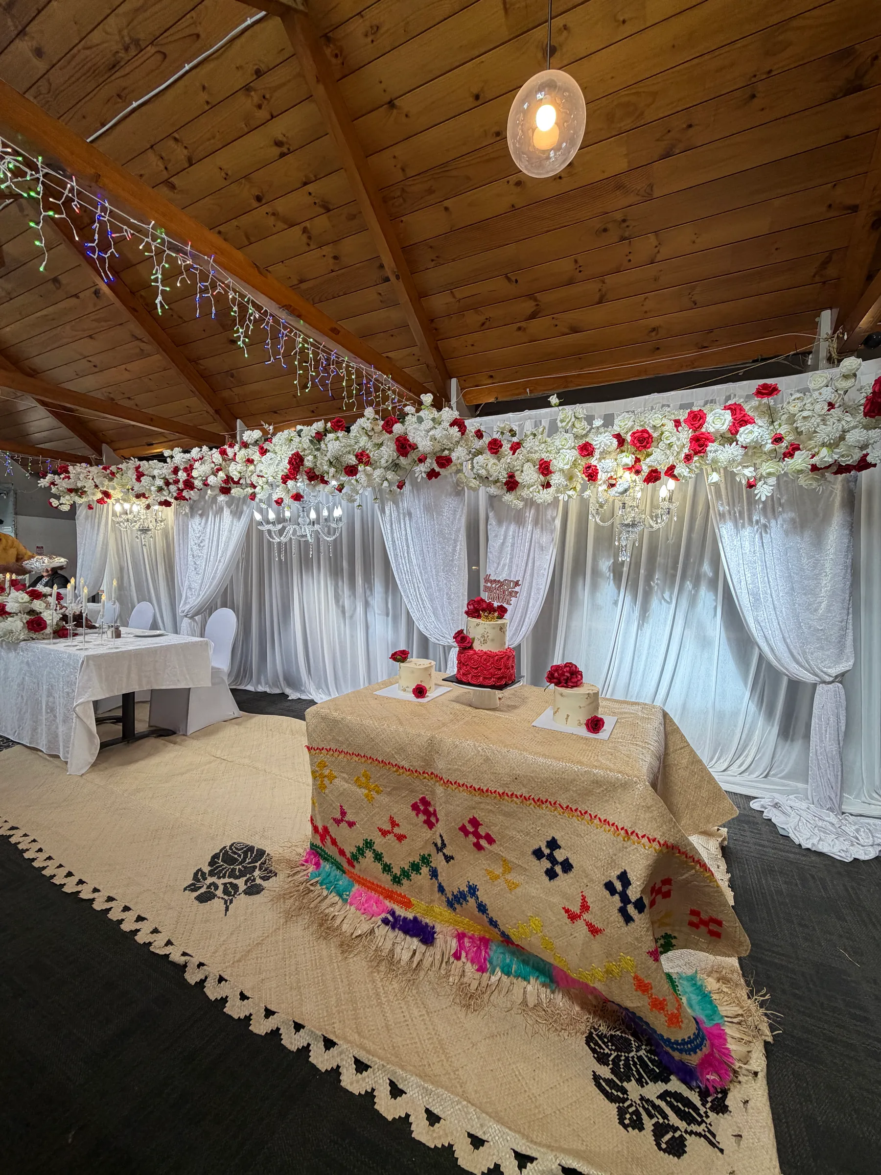 Birthday styling by Decoration X: Cake table draped in a woven mat with a bright stitched border and wool fringe, three cakes topped with red roses before a rose garland and white drapes.