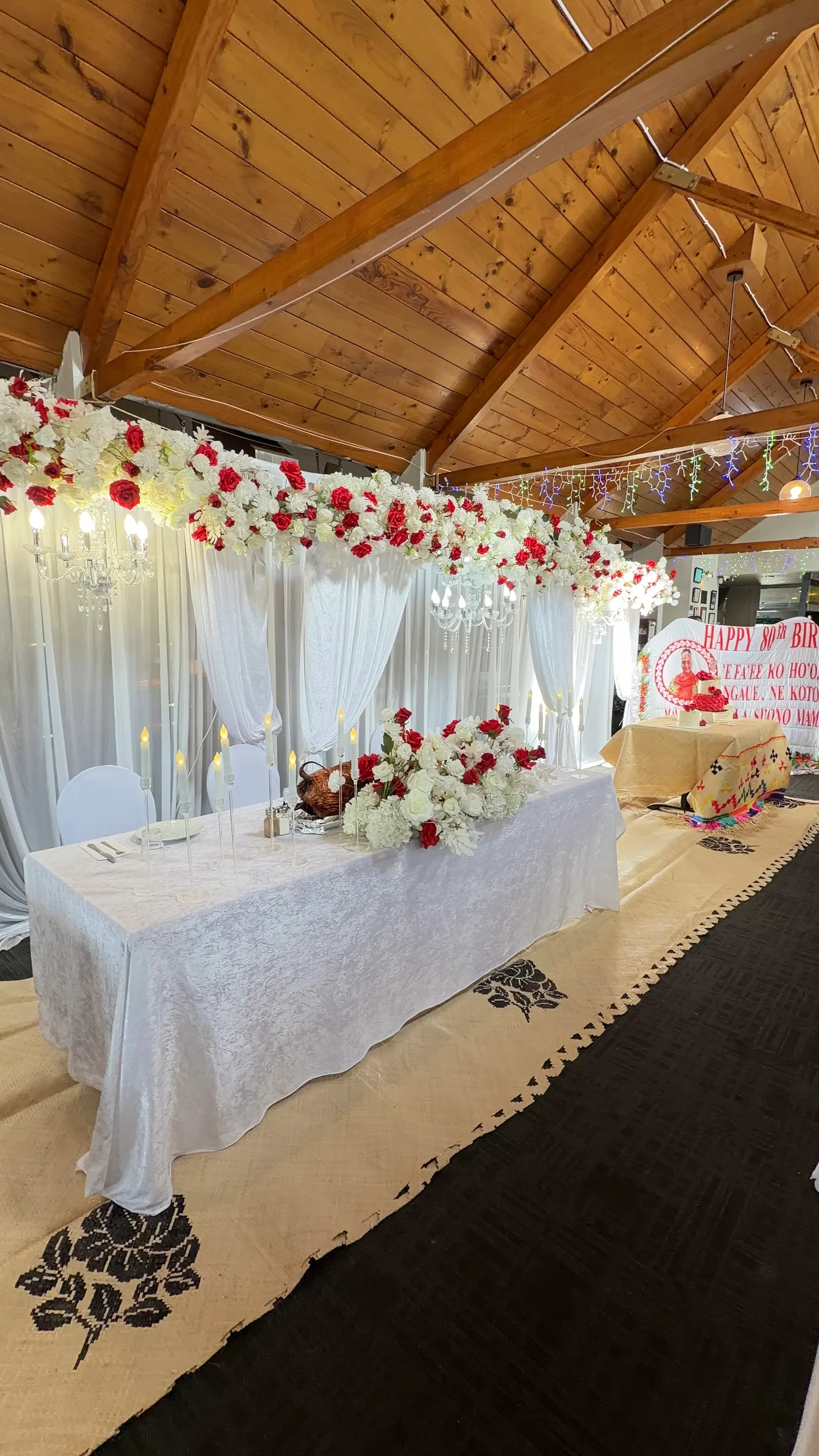 Birthday styling by Decoration X: Head table in white crushed velvet beneath a long red and white rose garland with crystal chandeliers, white drapes and patterned pandanus mats on the floor.