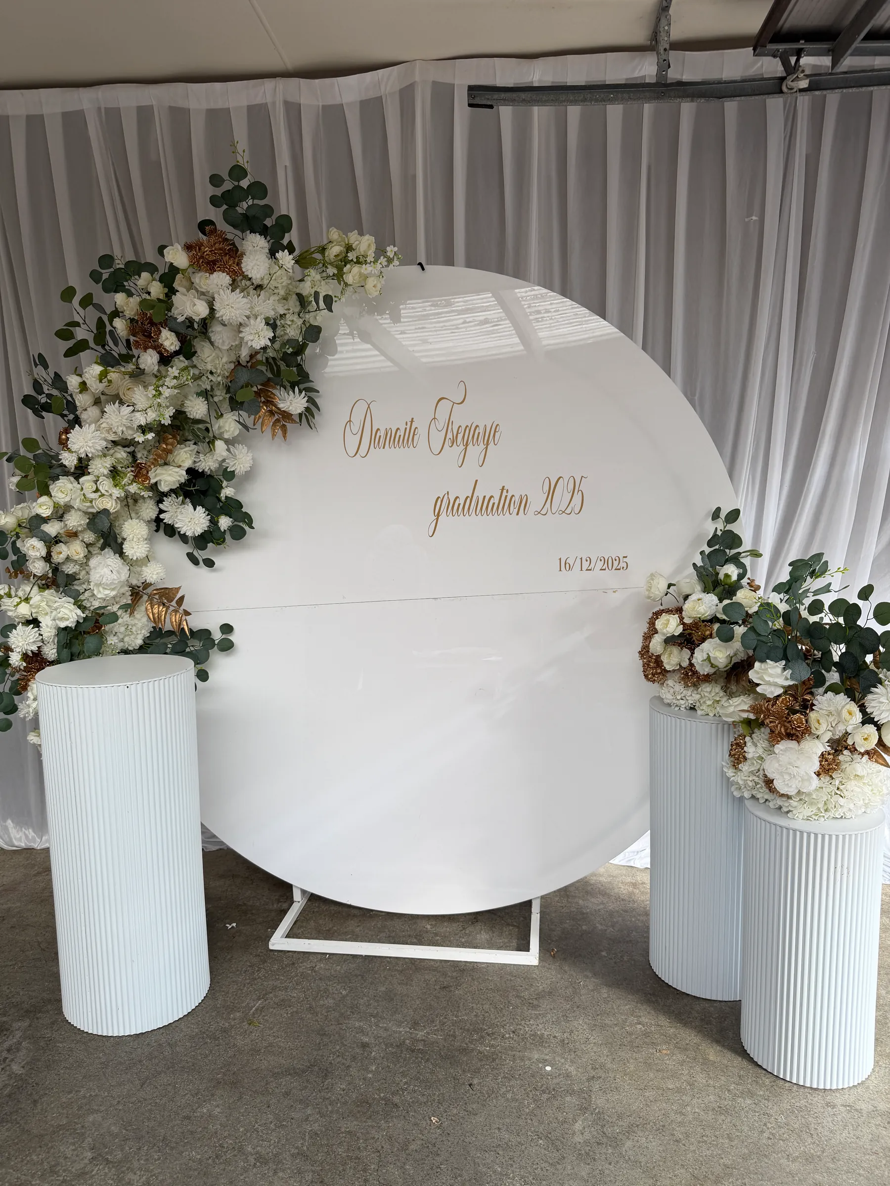 Graduation styling by Decoration X: White round disc with gold graduation lettering, a white and bronze rose and eucalyptus spray, and three fluted white plinths, two topped with florals.