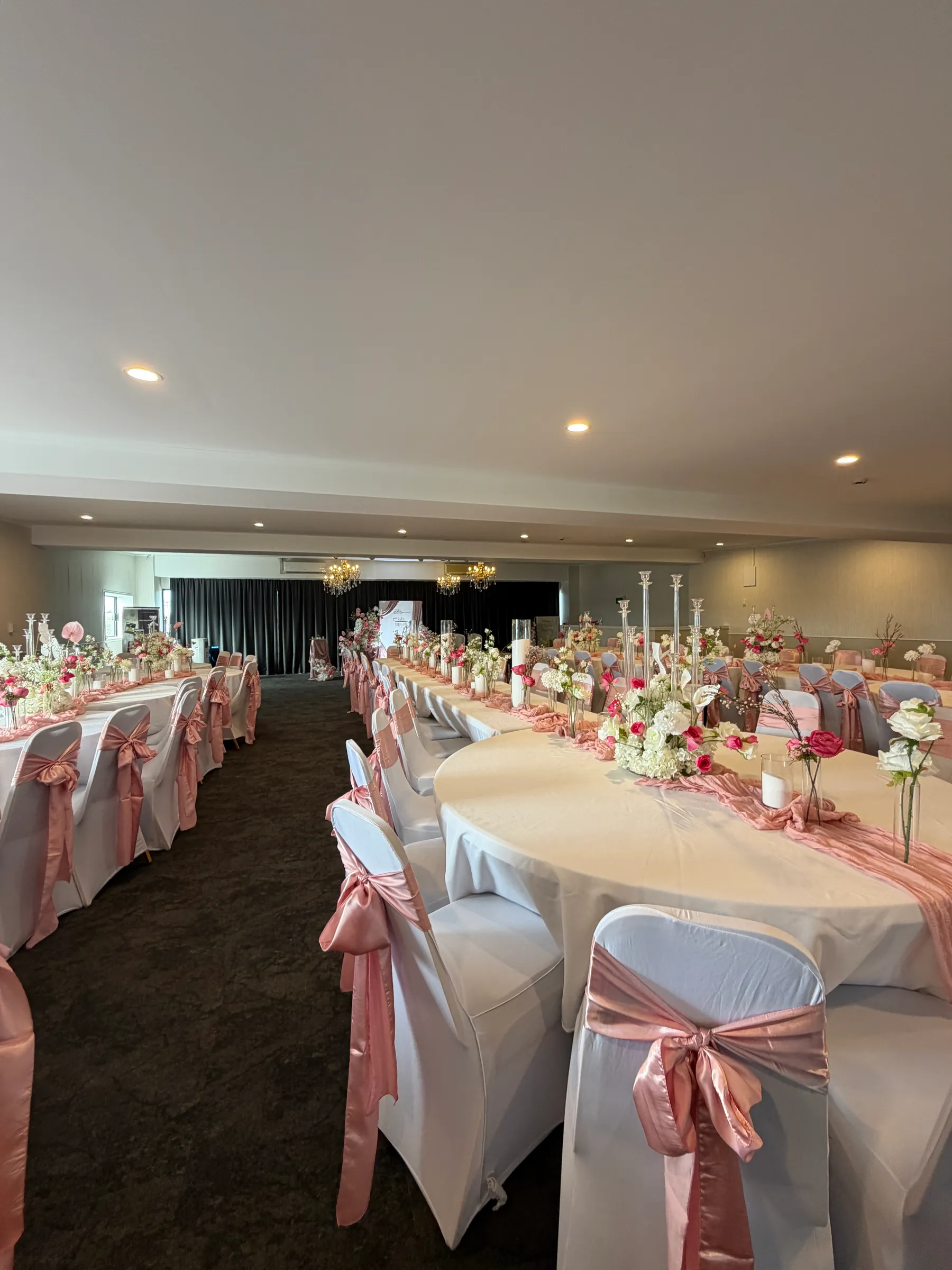 Reception styling by Decoration X: Long tables and a round table in white with blush satin chair sashes and runners, pink and white centrepieces and glass candelabra, black stage curtain beyond.