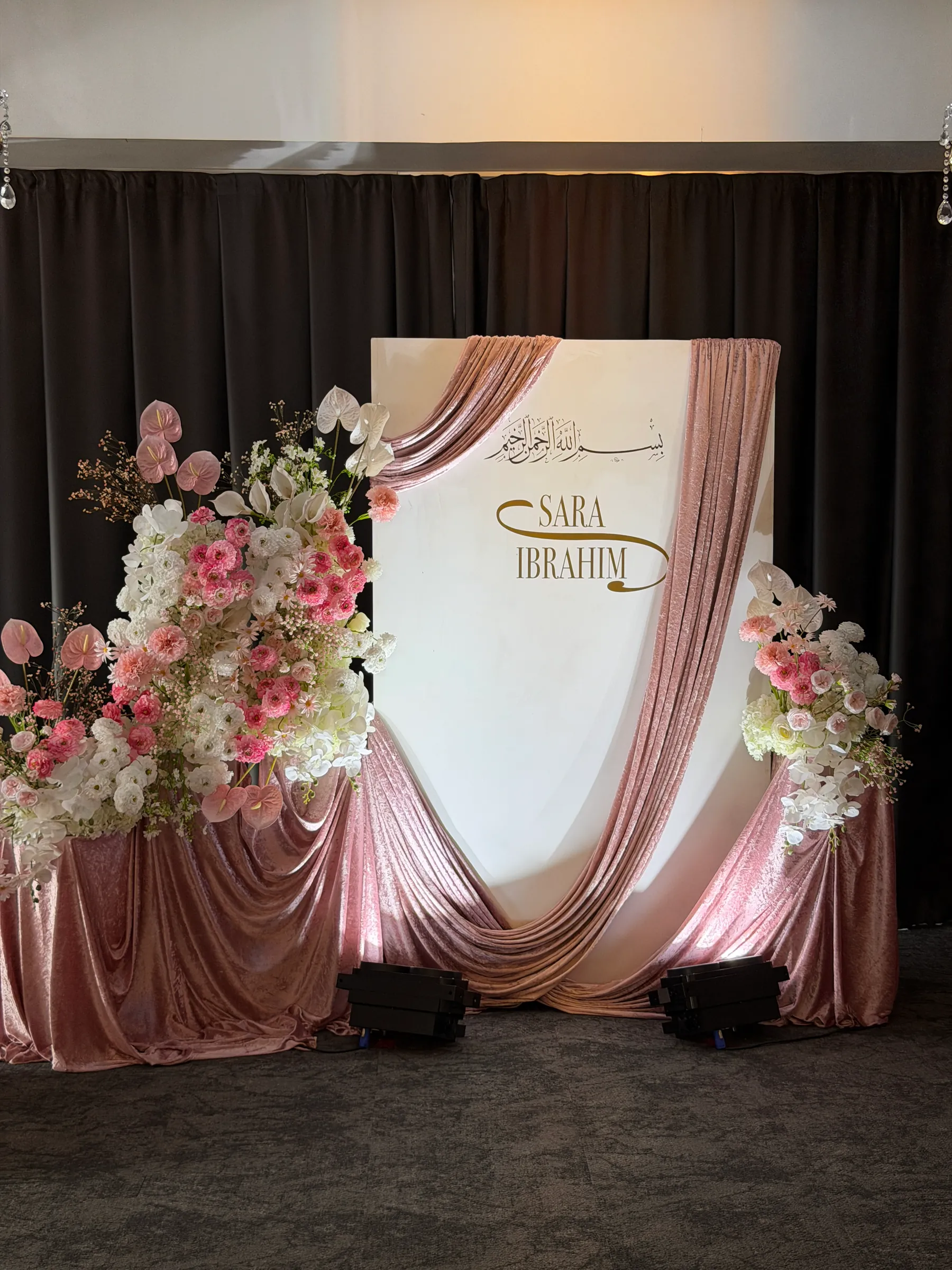 Stage styling by Decoration X: White panel backdrop with gold Arabic calligraphy and lettering, blush velvet swags, and pink and white anthurium, carnation and orchid sprays on draped plinths.