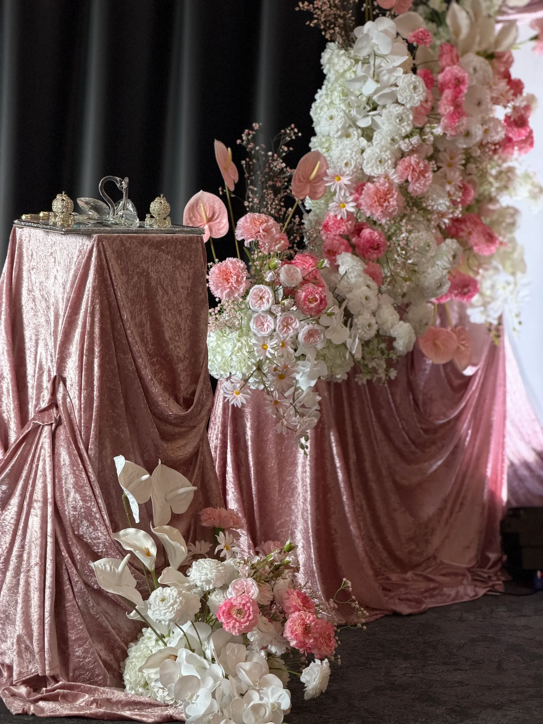 Stage styling by Decoration X: Blush crushed-velvet draped plinths with a glass swan and gold trinket boxes, beside tall cascades of pink carnations, white hydrangea, orchids and anthuriums.