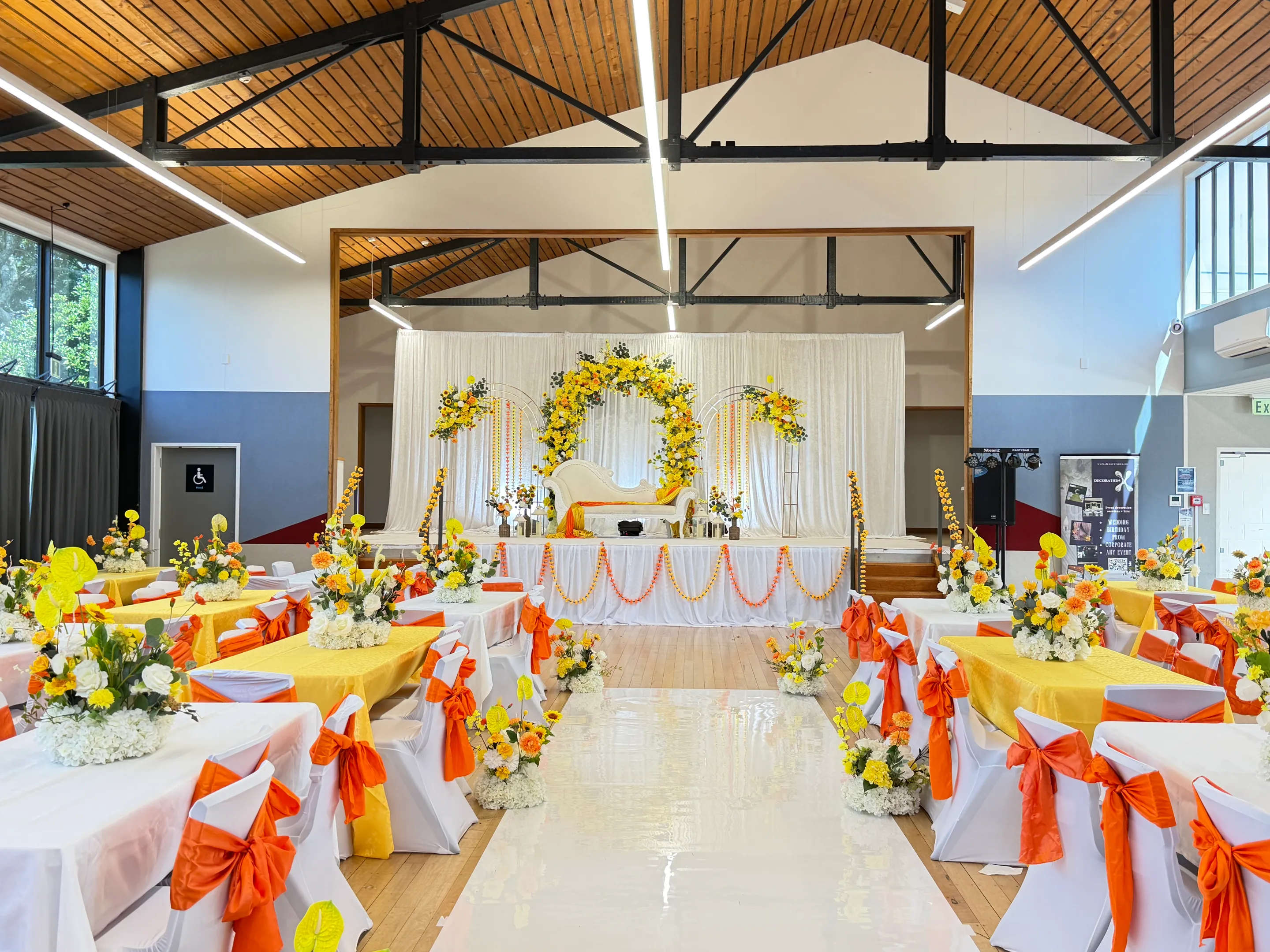 Cultural styling by Decoration X: Hall with a gloss white aisle runner between floral pedestals, tables in white and yellow with orange chair sashes, leading to a yellow and orange floral stage.