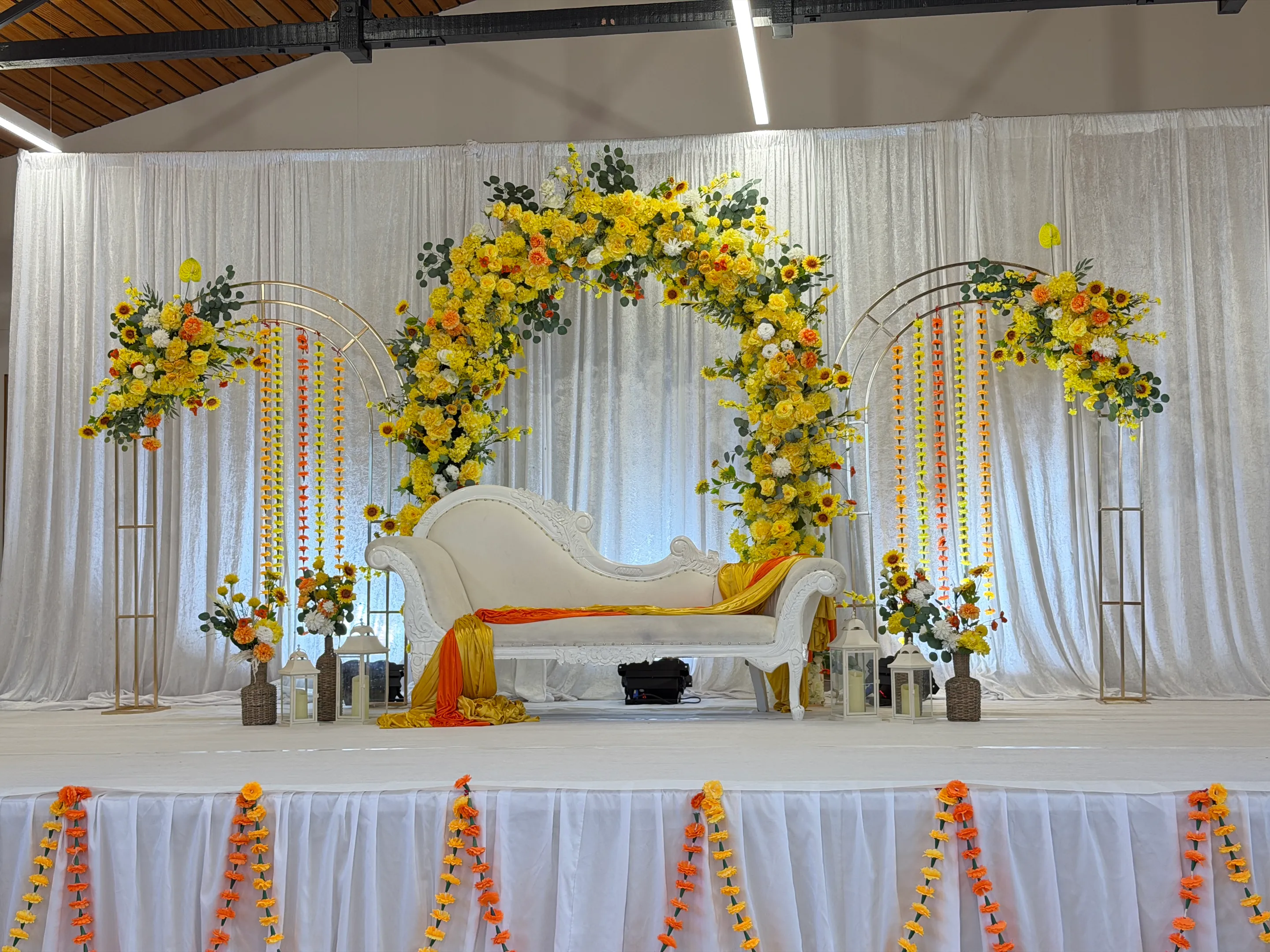 Cultural styling by Decoration X: White carved chaise draped in orange and yellow satin beneath a yellow and orange floral arch, gold hoop arches strung with marigolds, lanterns and white velvet drapes.