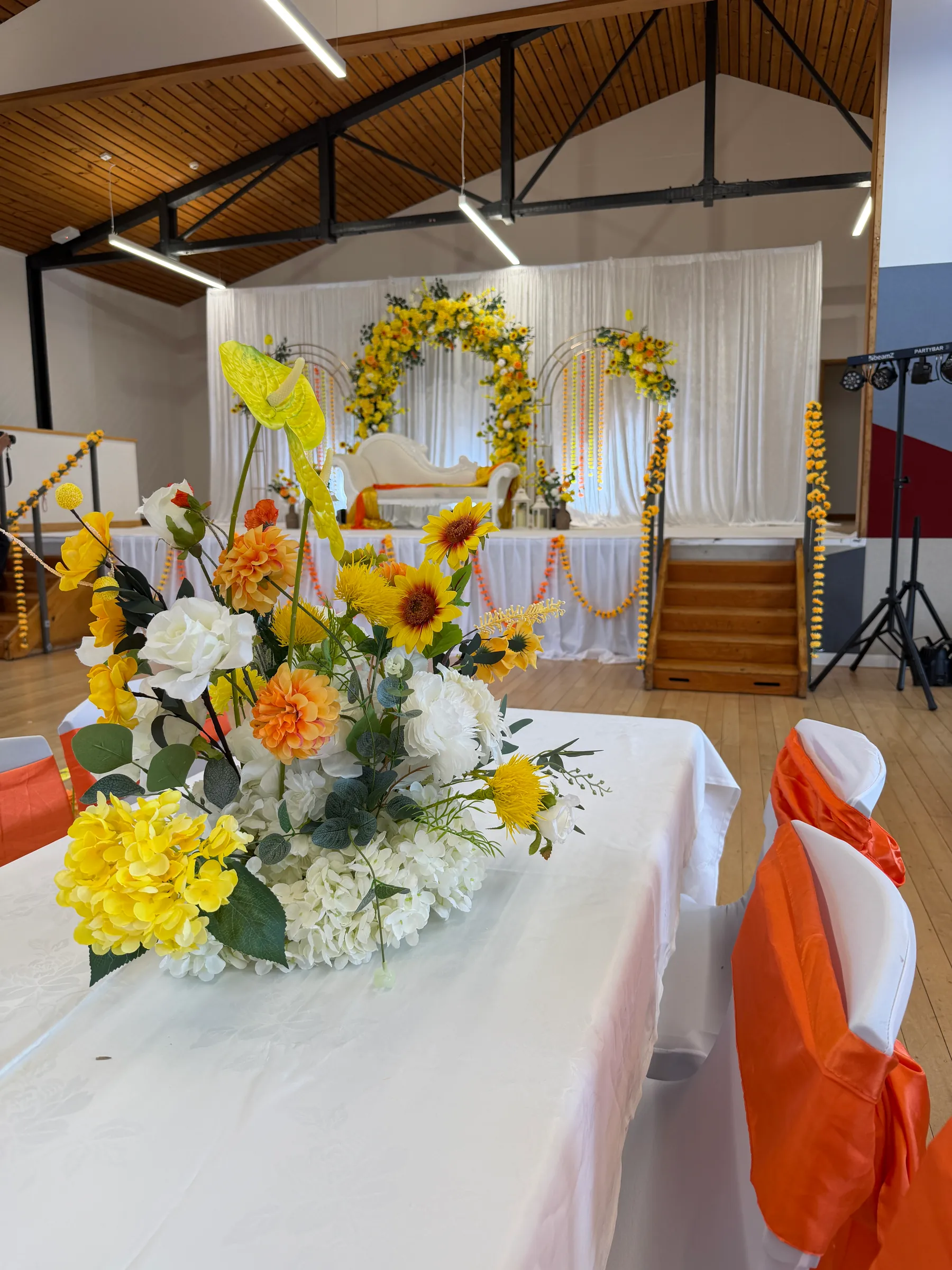Cultural styling by Decoration X: Centrepiece of sunflowers, orange dahlias, white hydrangea and a yellow anthurium on a white table, orange chair sashes and a floral stage beyond.