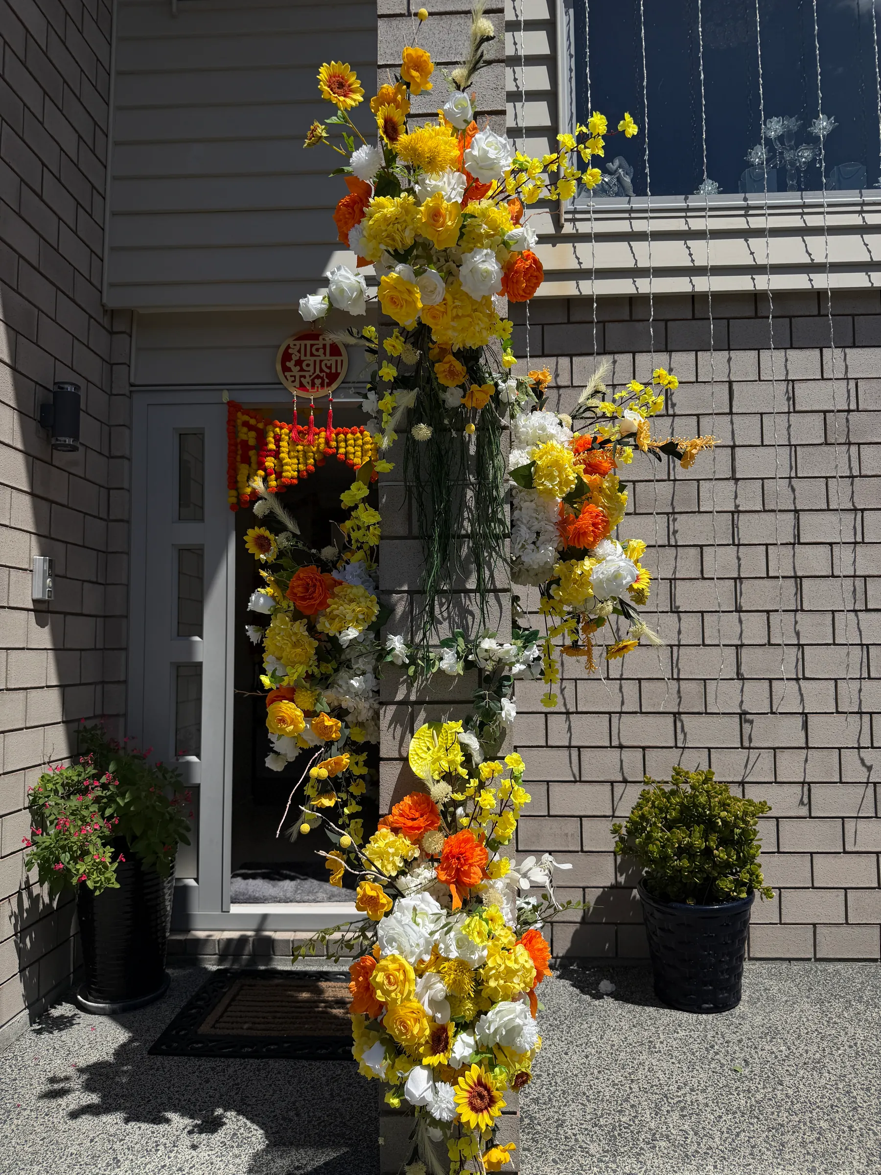 Cultural styling by Decoration X: Yellow, orange and white rose and sunflower cascade climbing a brick pillar at a front door hung with a marigold garland and a round Hindi sign.