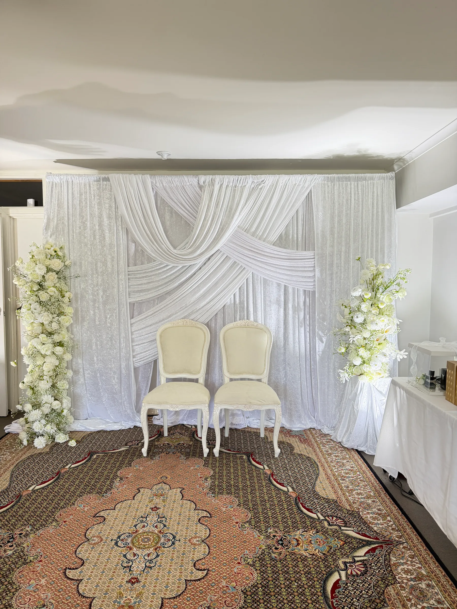Stage styling by Decoration X: White crushed-velvet backdrop with crossed swags behind two cream French chairs, tall white rose and gypsophila columns either side, on a Persian rug.