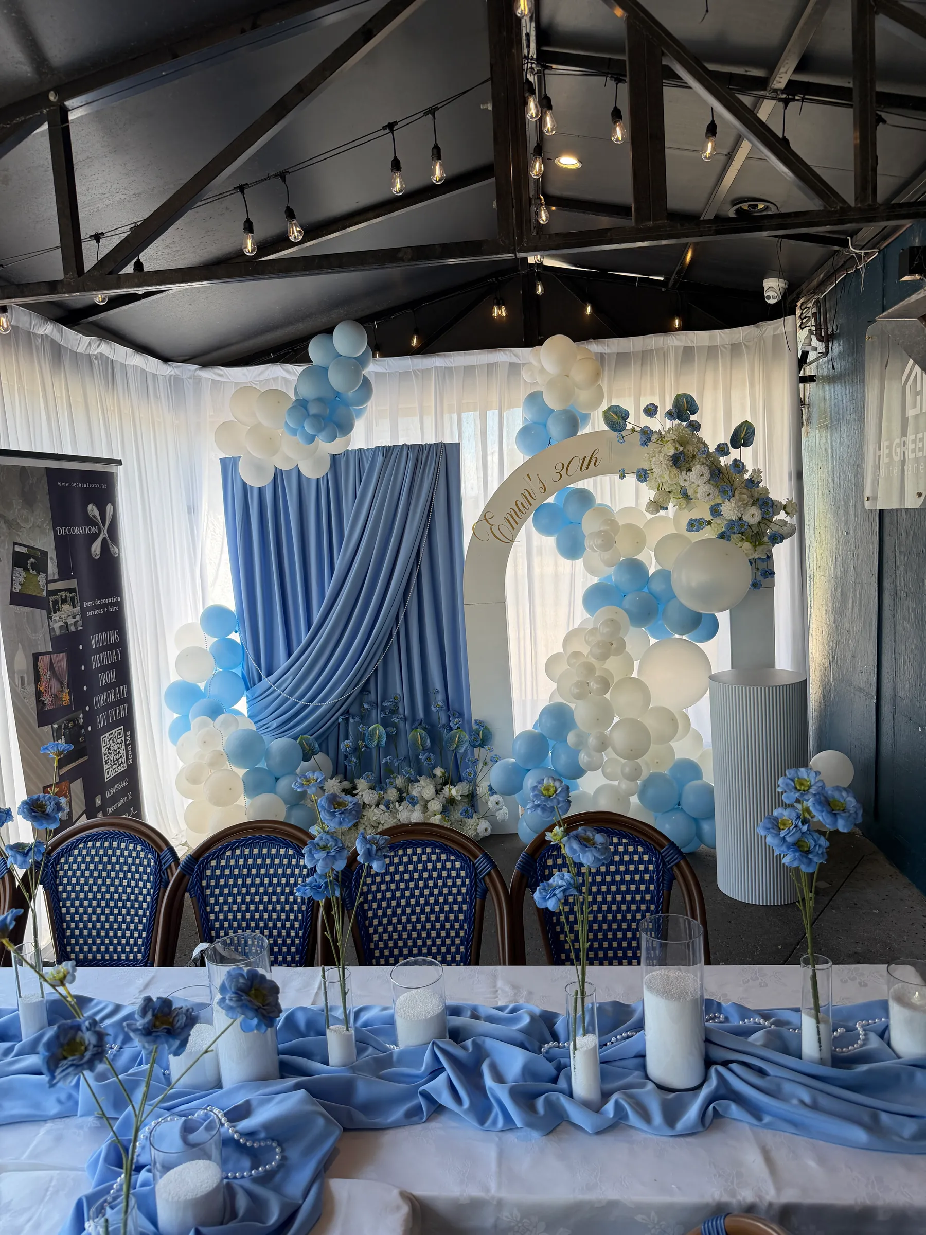 Birthday styling by Decoration X: Blue swagged drape and blue and white balloon garlands around a white name arch with blue and white florals, behind a head table of blue poppies and sand-filled vases.
