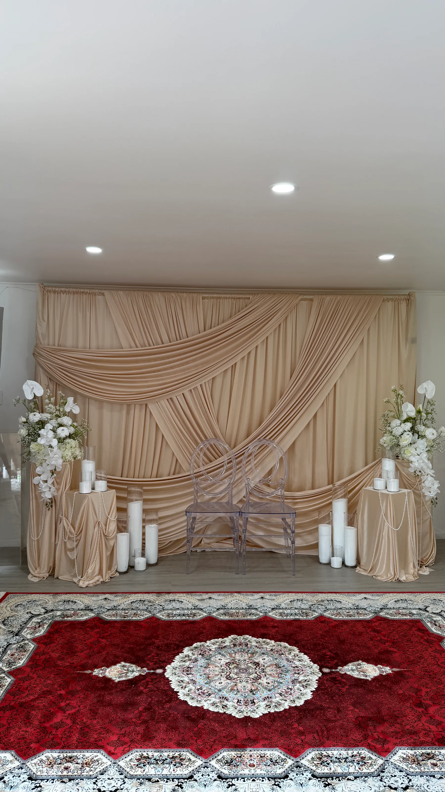 Stage styling by Decoration X: Champagne swagged drape behind two clear ghost chairs, white orchid and anthurium sprays on draped plinths, pillar candles, pearl strands and a red Persian rug.