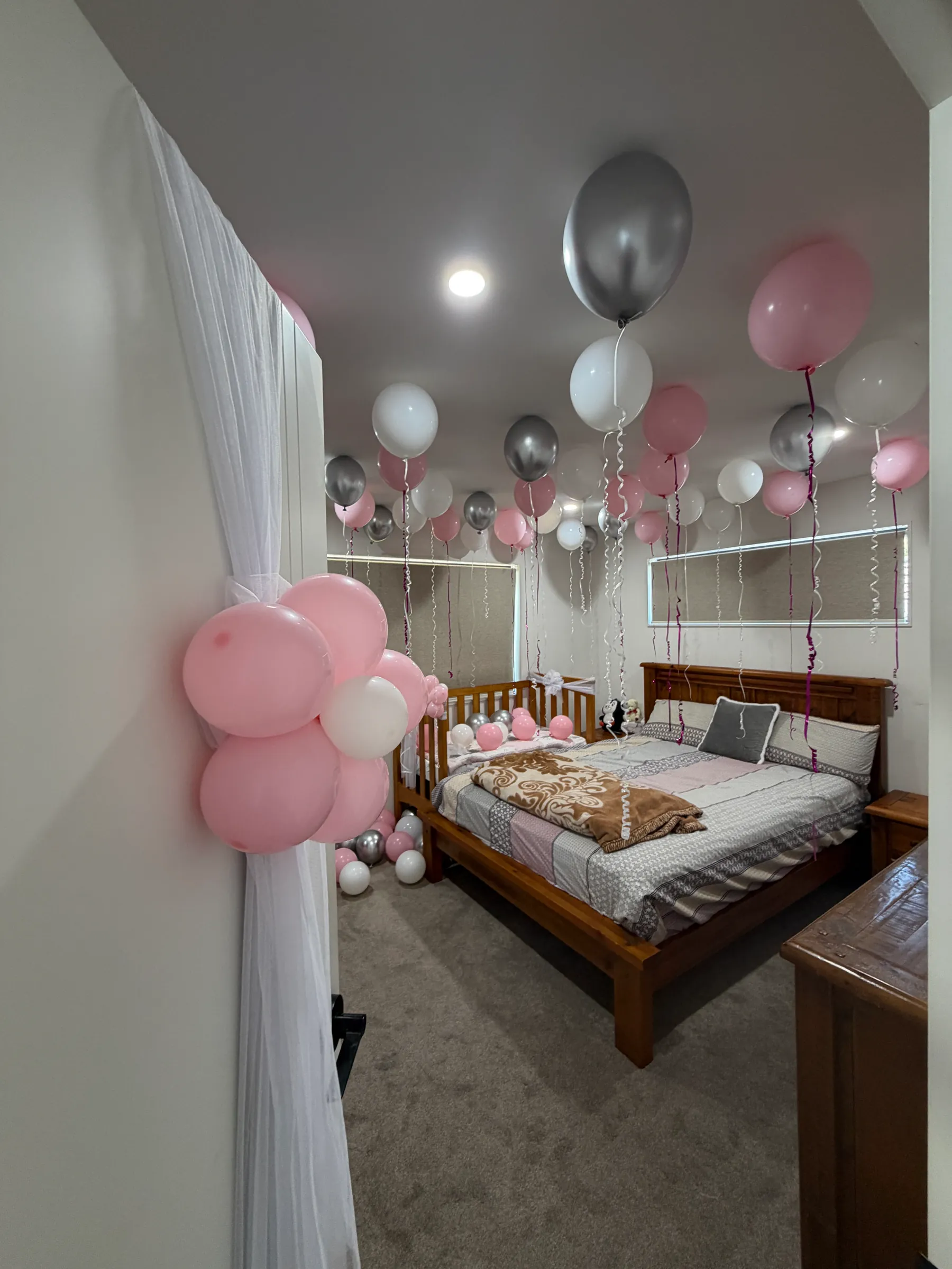 Baby shower styling by Decoration X: Pink, white and silver helium balloons float above a double bed and cot, a pink balloon cluster tied to a sheer white curtain at the doorway.