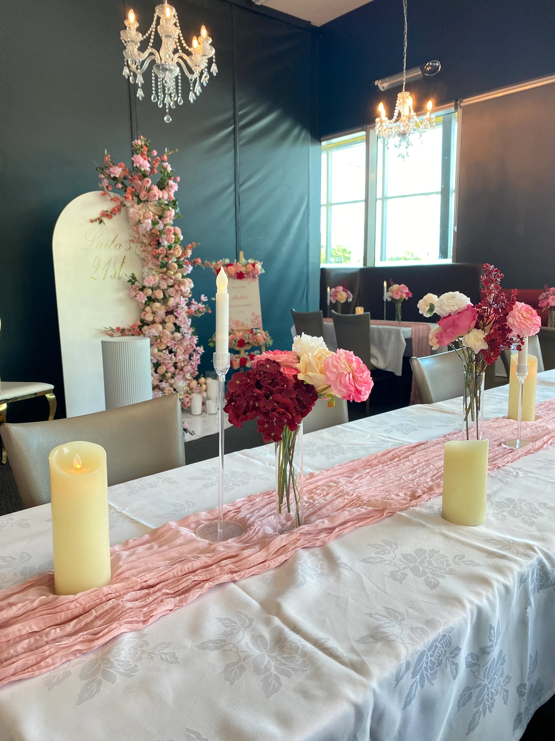 Birthday styling by Decoration X: Table set with a pink cheesecloth runner, burgundy and pink posies and LED candles, before a white arch name board with a pink rose cascade and a floral easel sign.