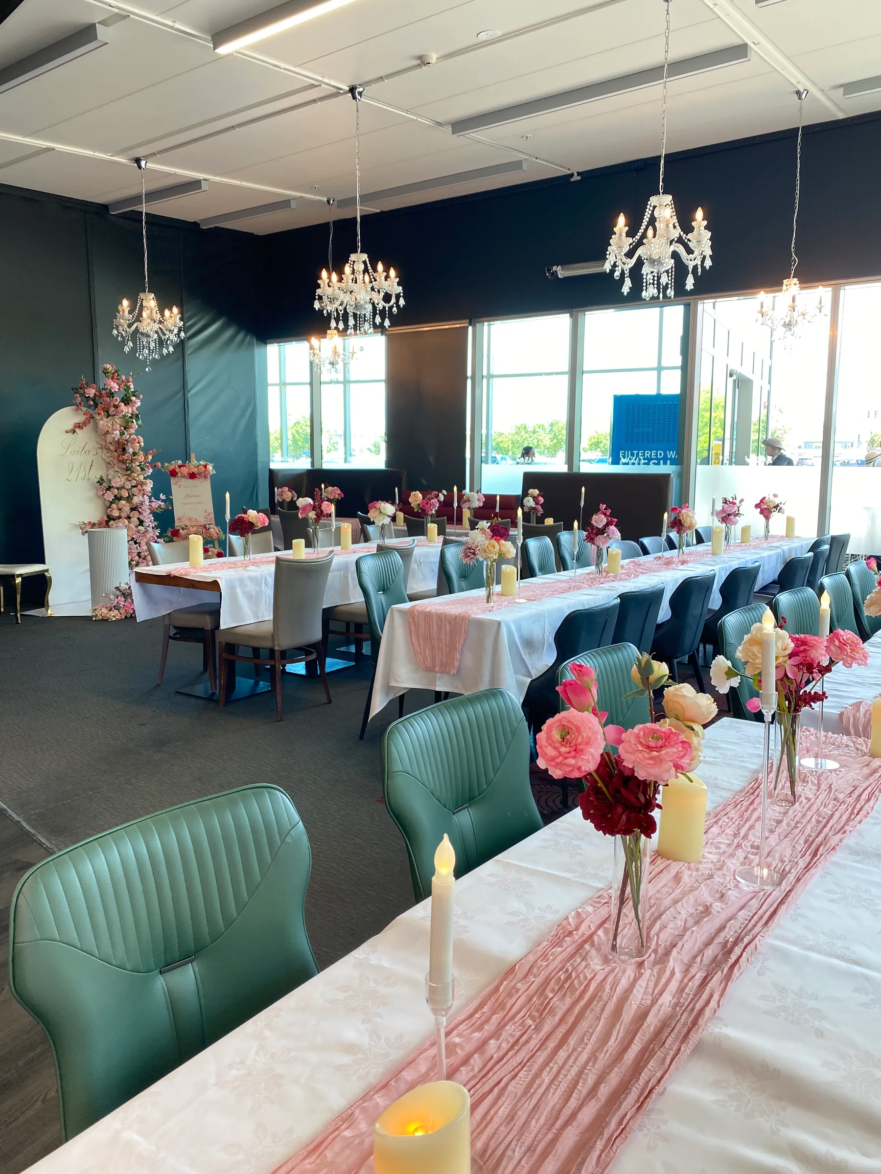 Birthday styling by Decoration X: Function room with chandeliers over long tables of white cloths, pink cheesecloth runners, rose posies and LED candles, teal chairs and a floral arch board beyond.