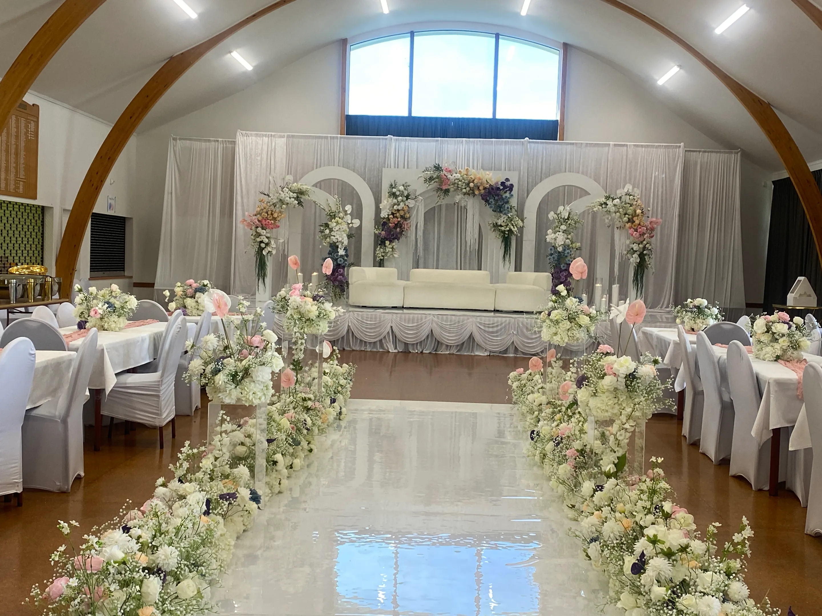 Reception styling by Decoration X: Hall with a glossy white aisle lined in white, pink and navy floral runners and plinths, leading past white-covered chairs and tables to a floral arch sofa stage.