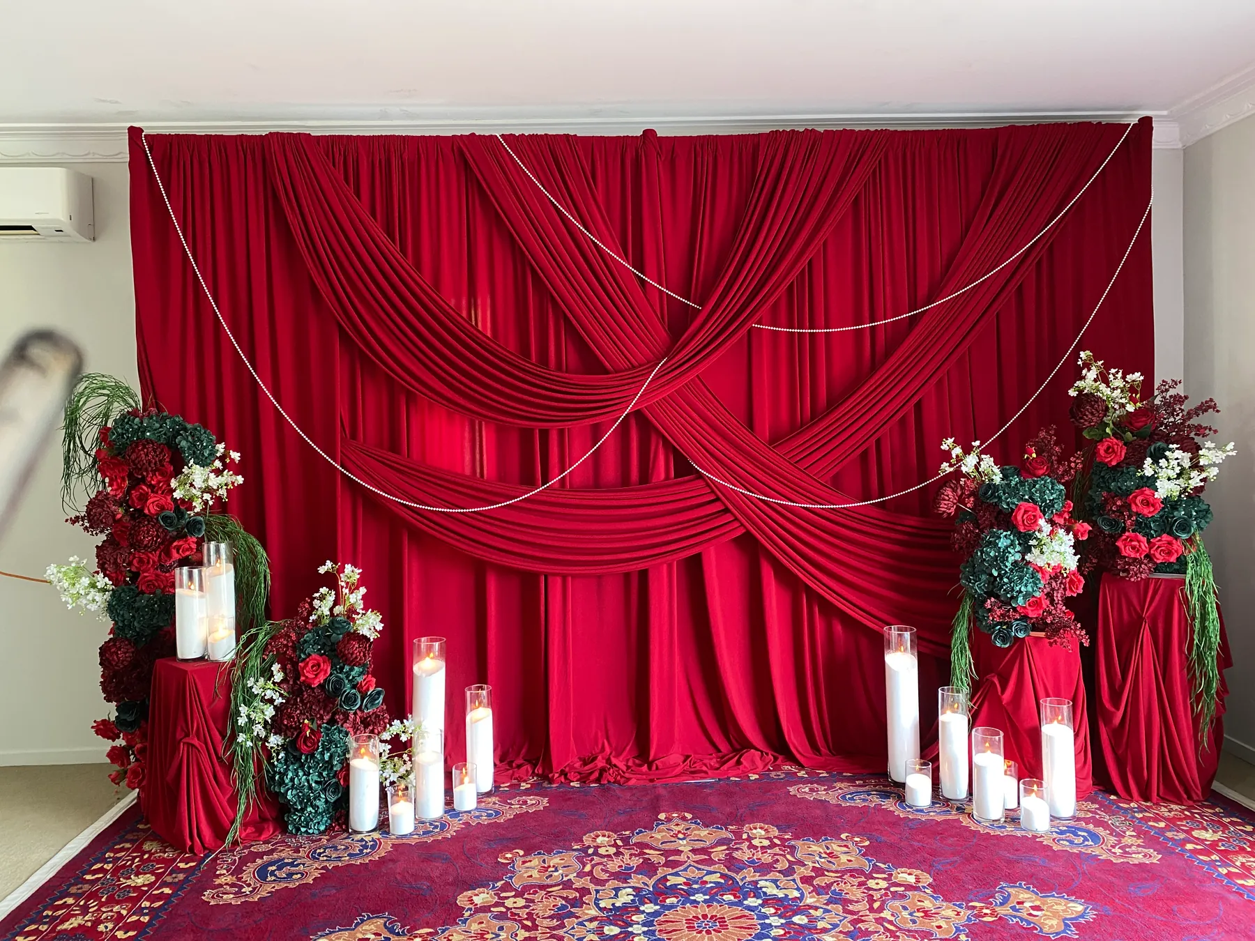 Stage styling by Decoration X: Deep red velvet backdrop with crossed swags and pearl strands, red-draped plinths of red roses and dark green hydrangea, and glass cylinder candles on a Persian rug.