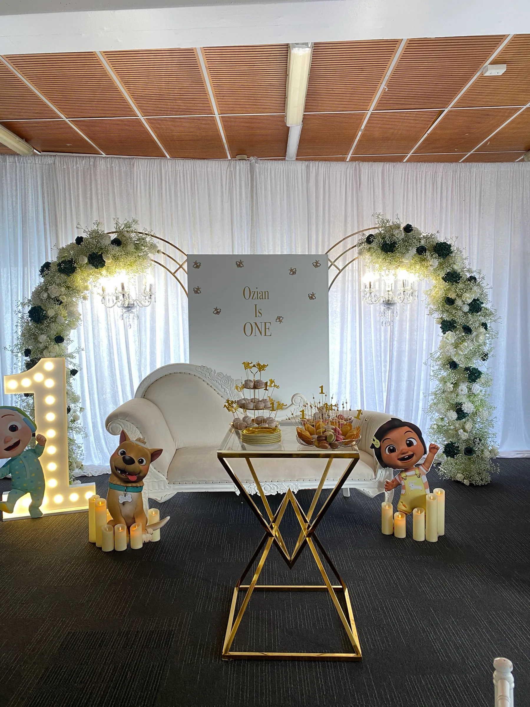 Birthday styling by Decoration X: Name board between two arches of white and dark green roses with chandeliers, a carved white sofa, a gold geometric dessert table, a marquee one and cut-outs.