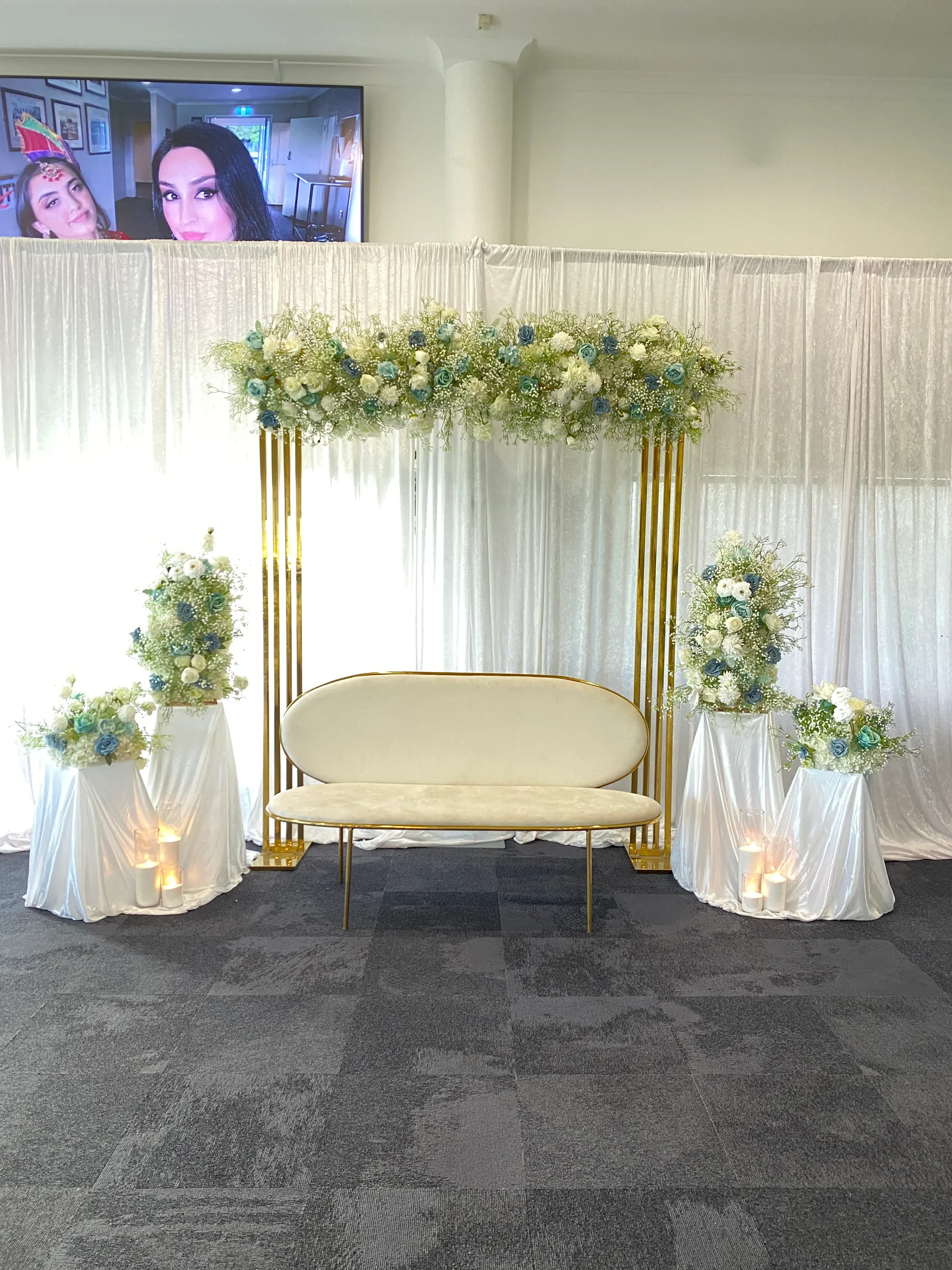Stage styling by Decoration X: Gold slatted arch topped with white roses, blue roses and gypsophila, a cream oval sofa, and white-draped plinths with matching florals and pillar candles.