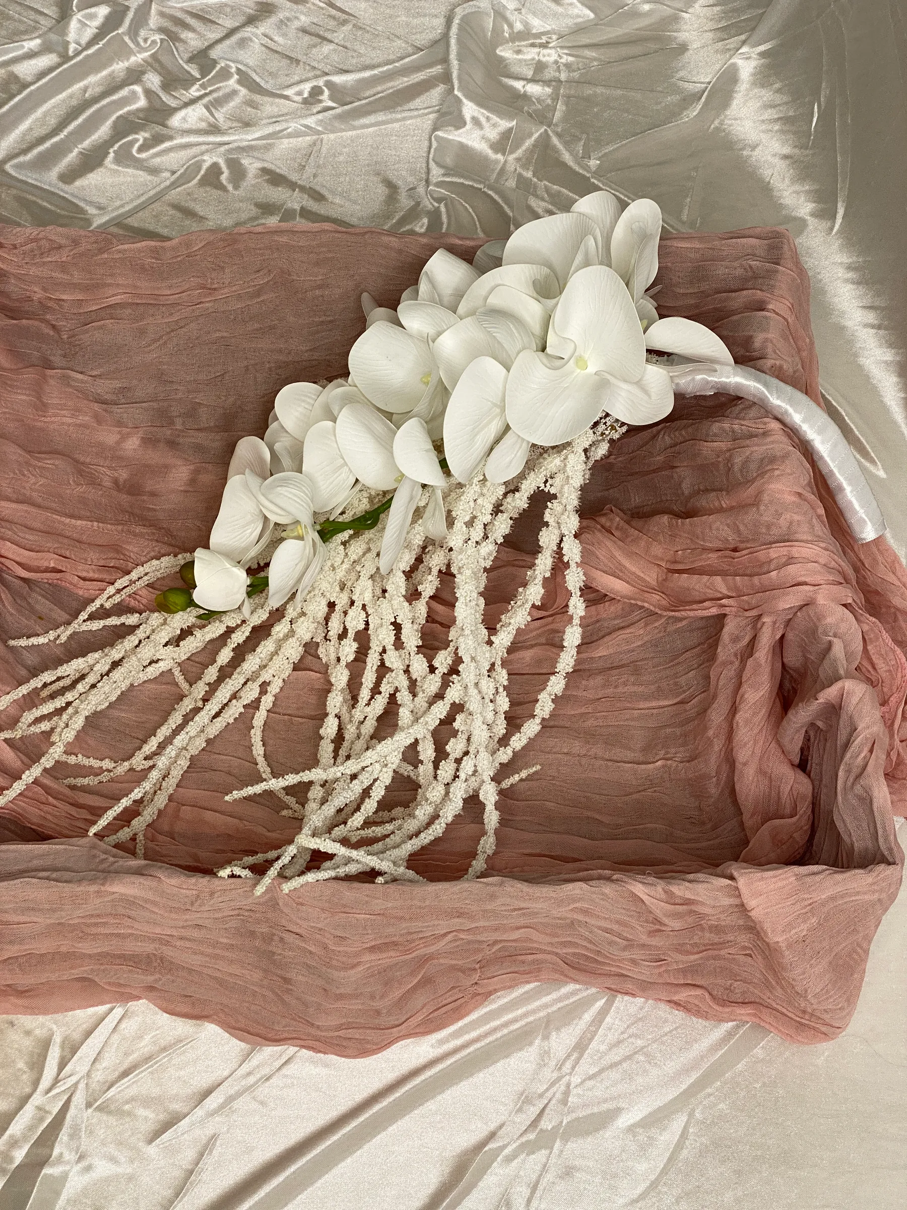 Ceremony styling by Decoration X: Cascading bridal bouquet of white phalaenopsis orchids with trailing white beaded amaranthus and a satin-wrapped handle, on dusty pink cheesecloth.