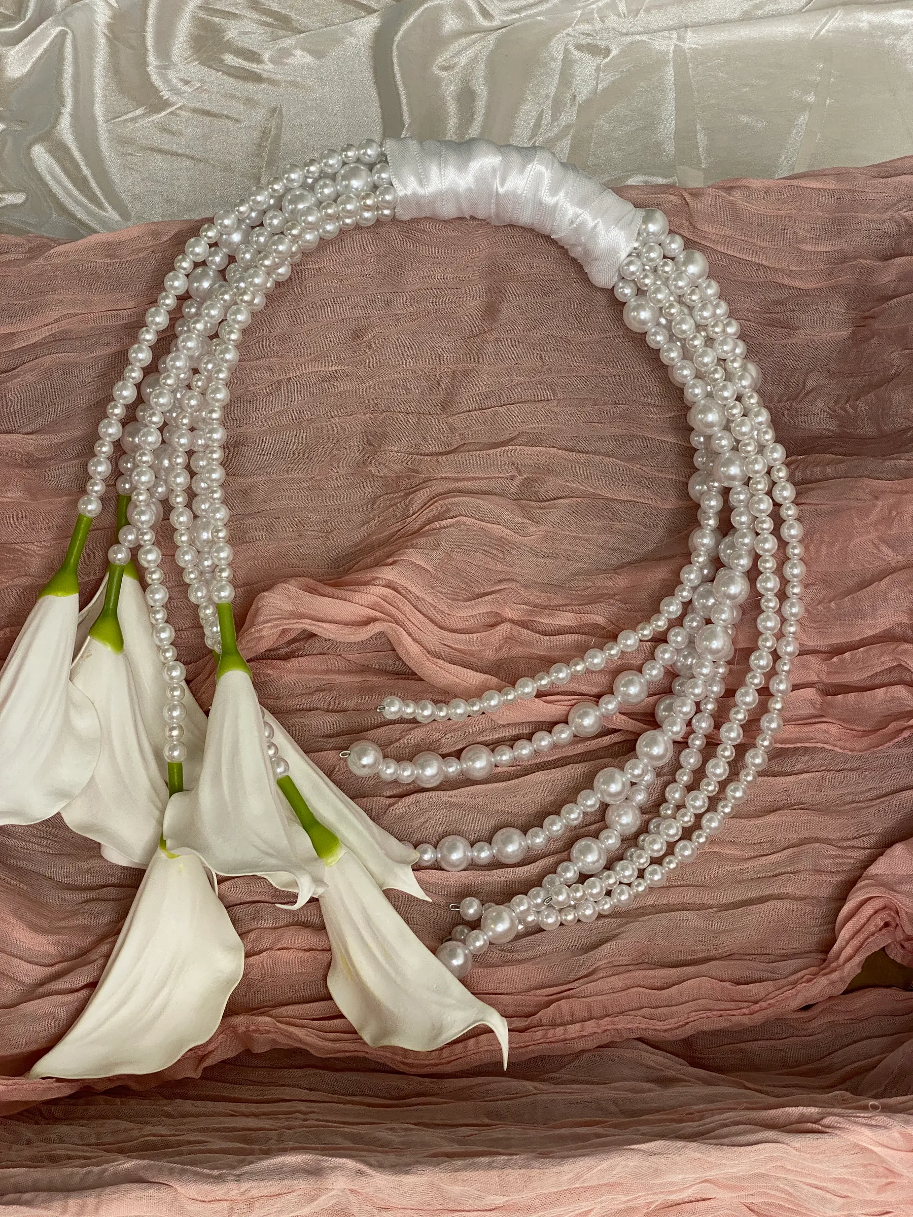 Ceremony styling by Decoration X: Bridal bouquet of looped pearl strands on a white satin-wrapped handle, white calla lilies trailing from one end, on dusty pink cheesecloth over white satin.