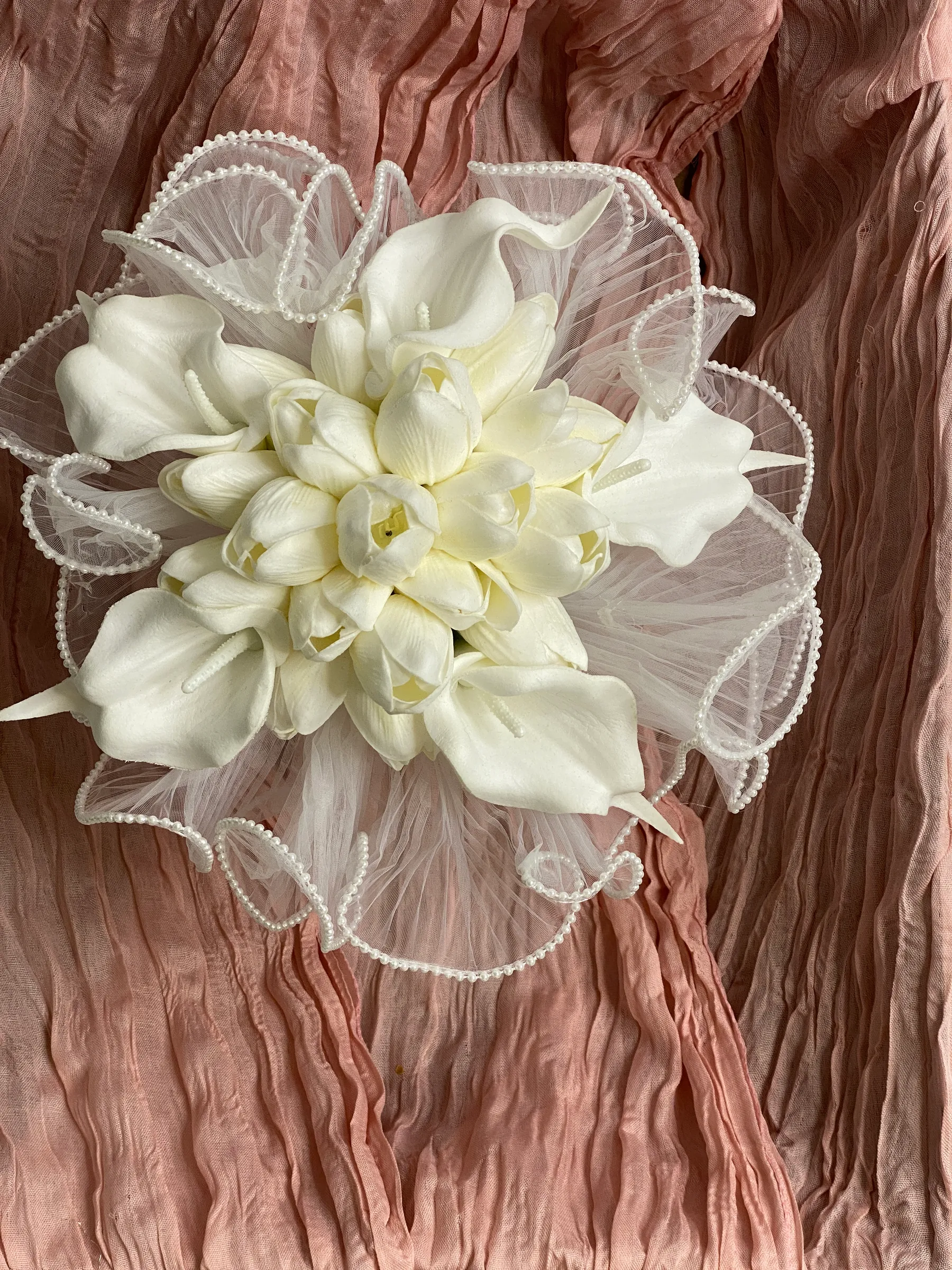 Ceremony styling by Decoration X: Overhead view of a white tulip and calla lily bridal bouquet ringed in pearl-edged pleated tulle, laid on dusty pink cheesecloth.