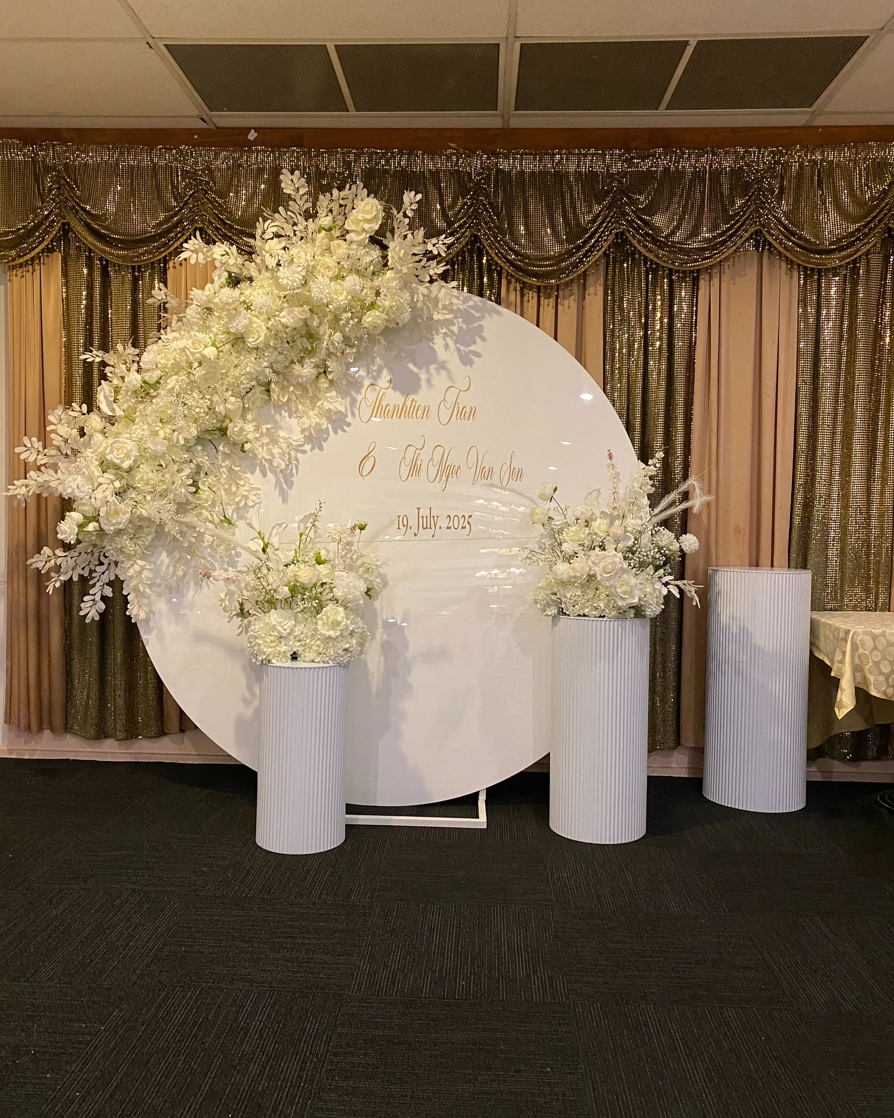 Stage styling by Decoration X: Round white backdrop with gold script names, white roses and foliage sweeping across its edge, and ribbed white plinths of white florals before gold curtains.