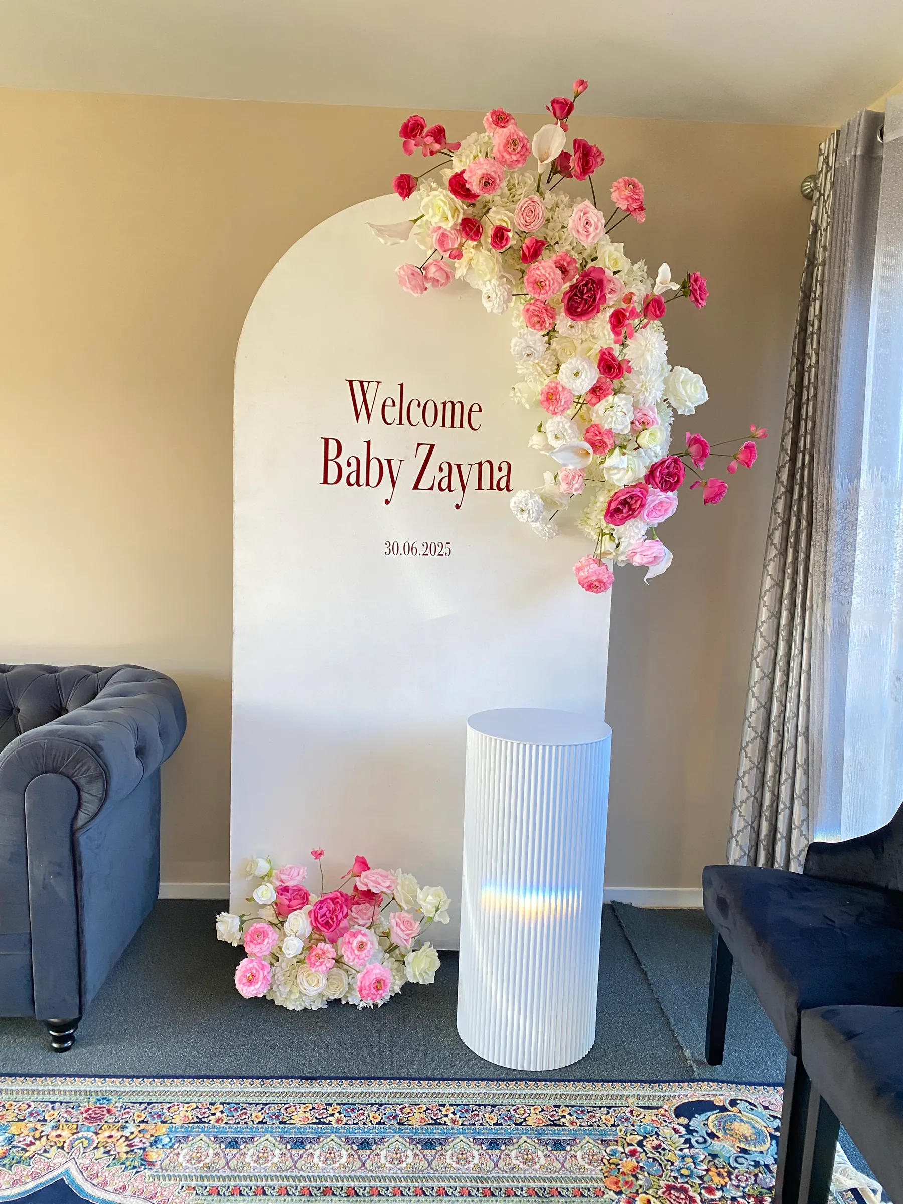 Baby shower styling by Decoration X: Cream arch board with a welcome-baby name in burgundy lettering, a cascade of pink and white roses, a ribbed white plinth and a floor posy in a living room.