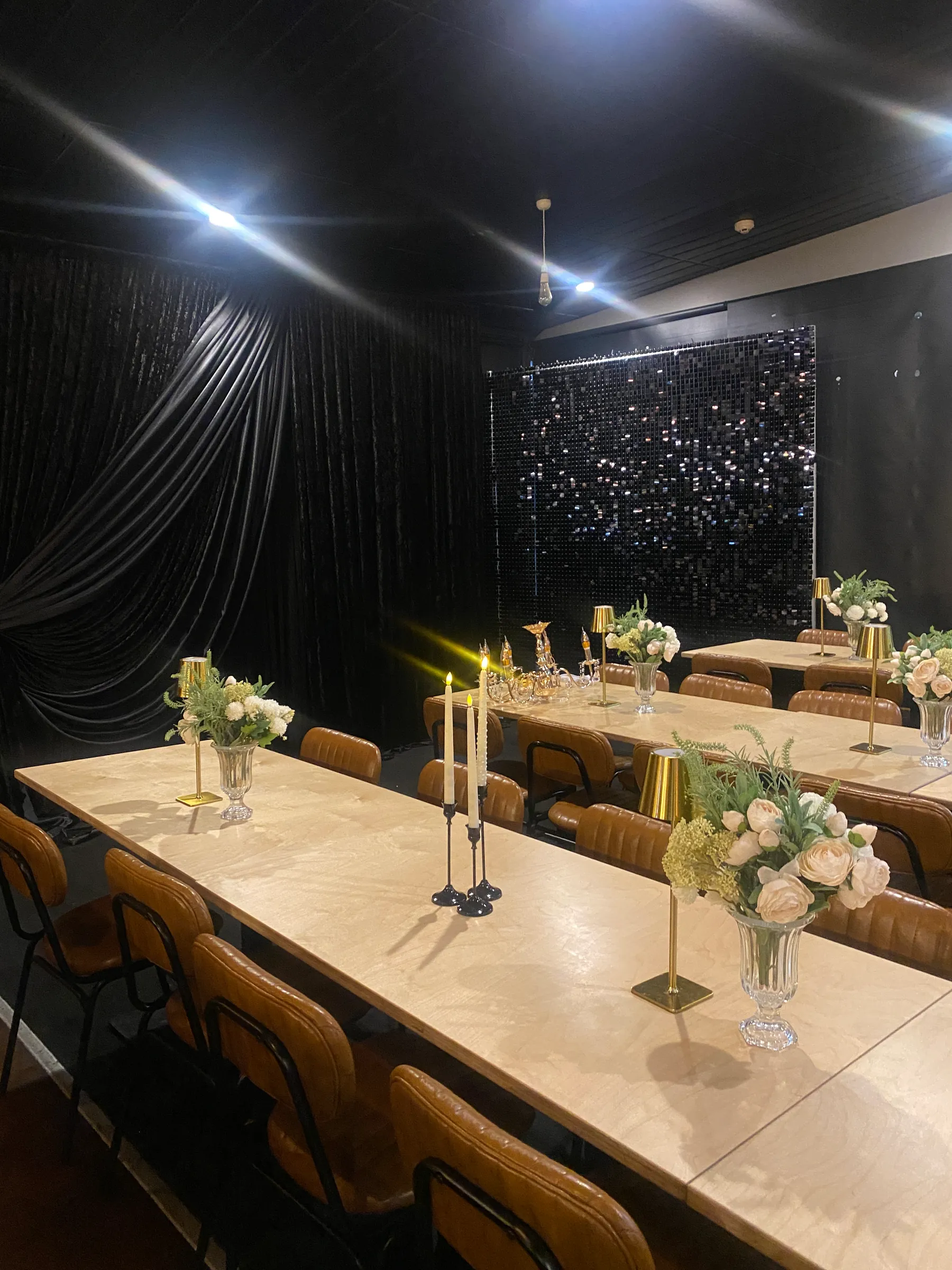 Reception styling by Decoration X: Long marble-look tables with tan leather chairs, gold lamps, taper candles and white rose posies before a black velvet drape and a black sequin shimmer wall.
