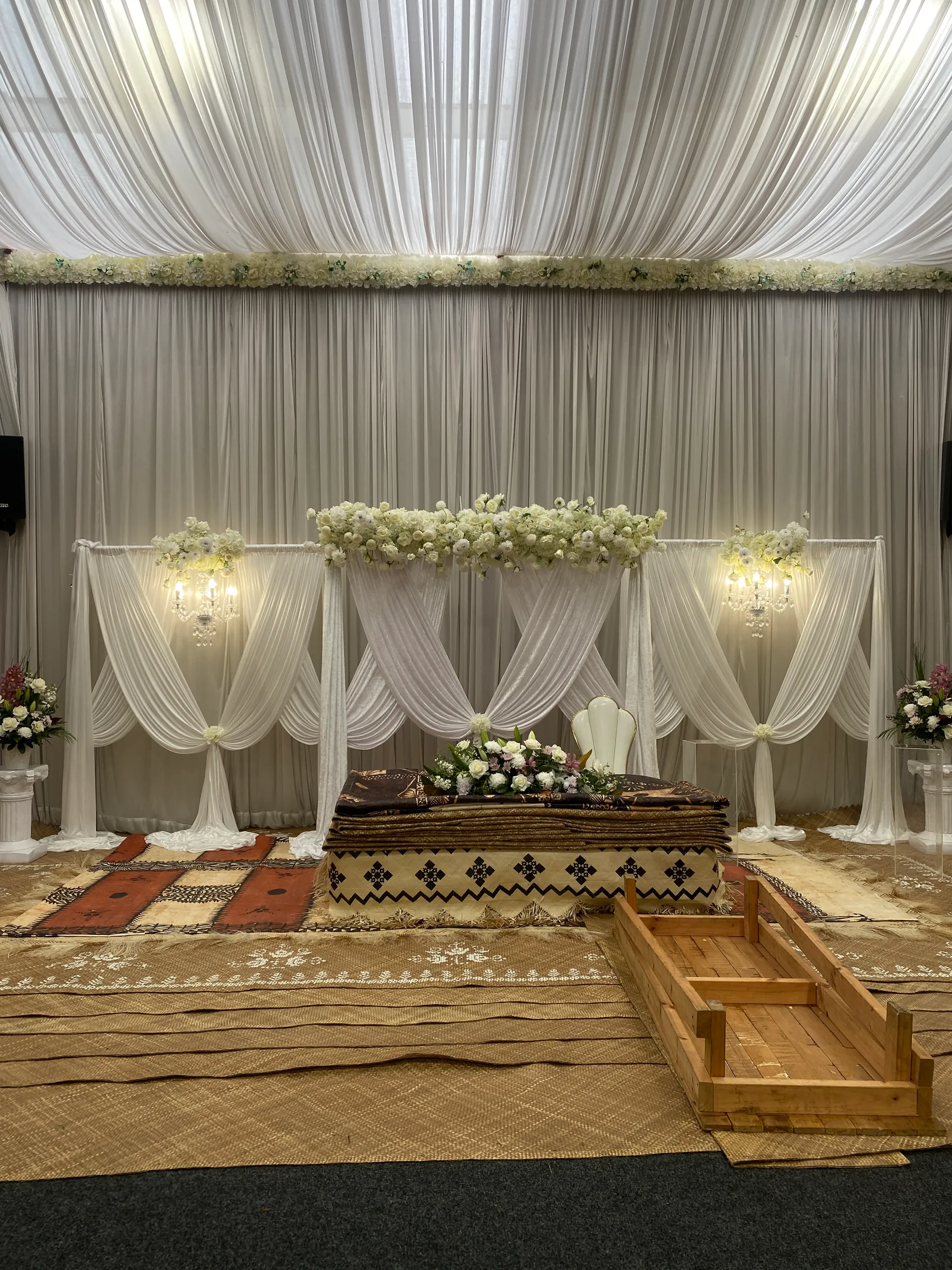 Cultural styling by Decoration X: White swagged drapes under a floral ceiling garland, a tapa and fine-mat platform topped with white roses, a white throne chair, woven mats and an open wooden frame.