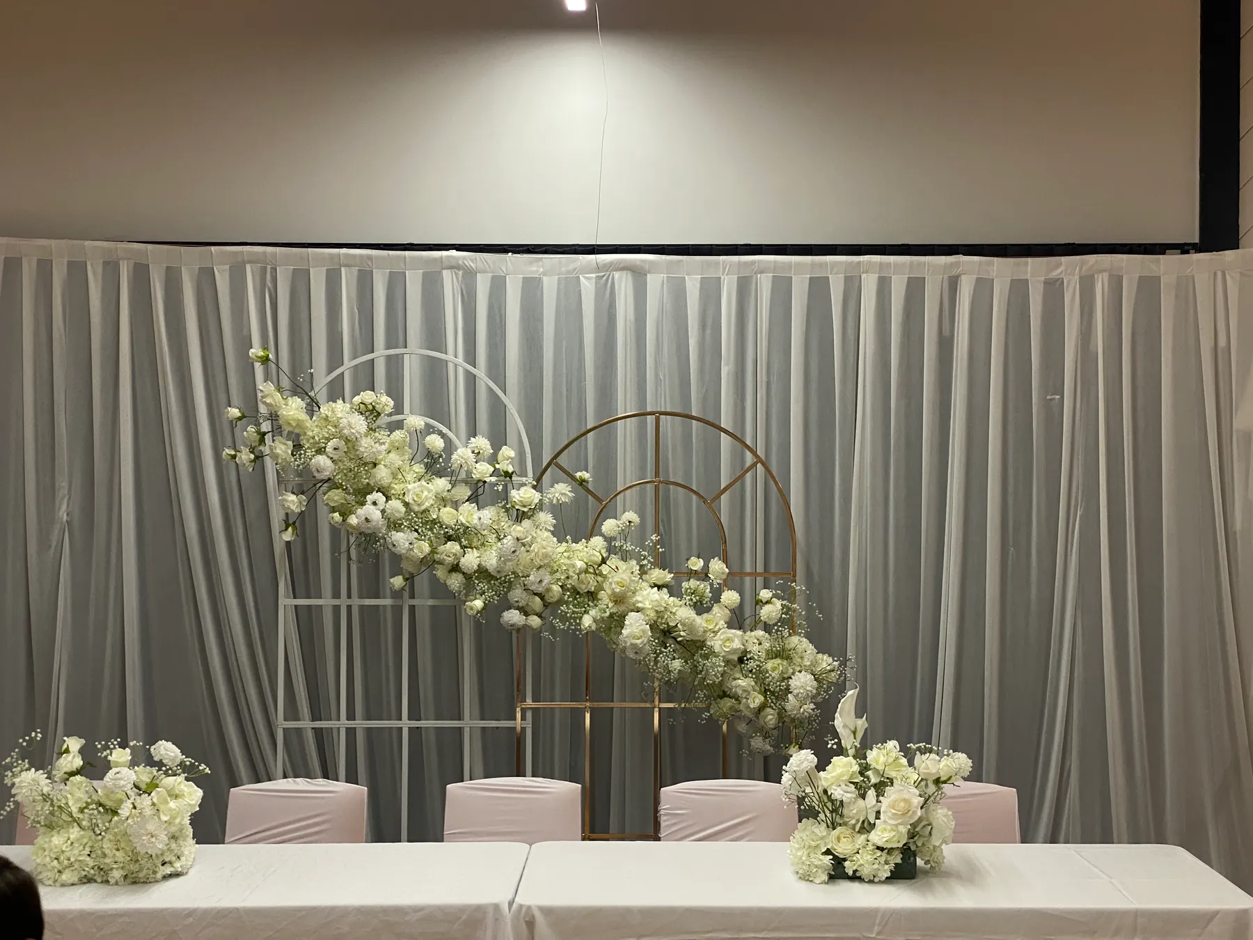 Stage styling by Decoration X: Head table with white floral mounds before white and gold grid arches joined by a cascade of white roses and gypsophila, pink-covered chairs behind.