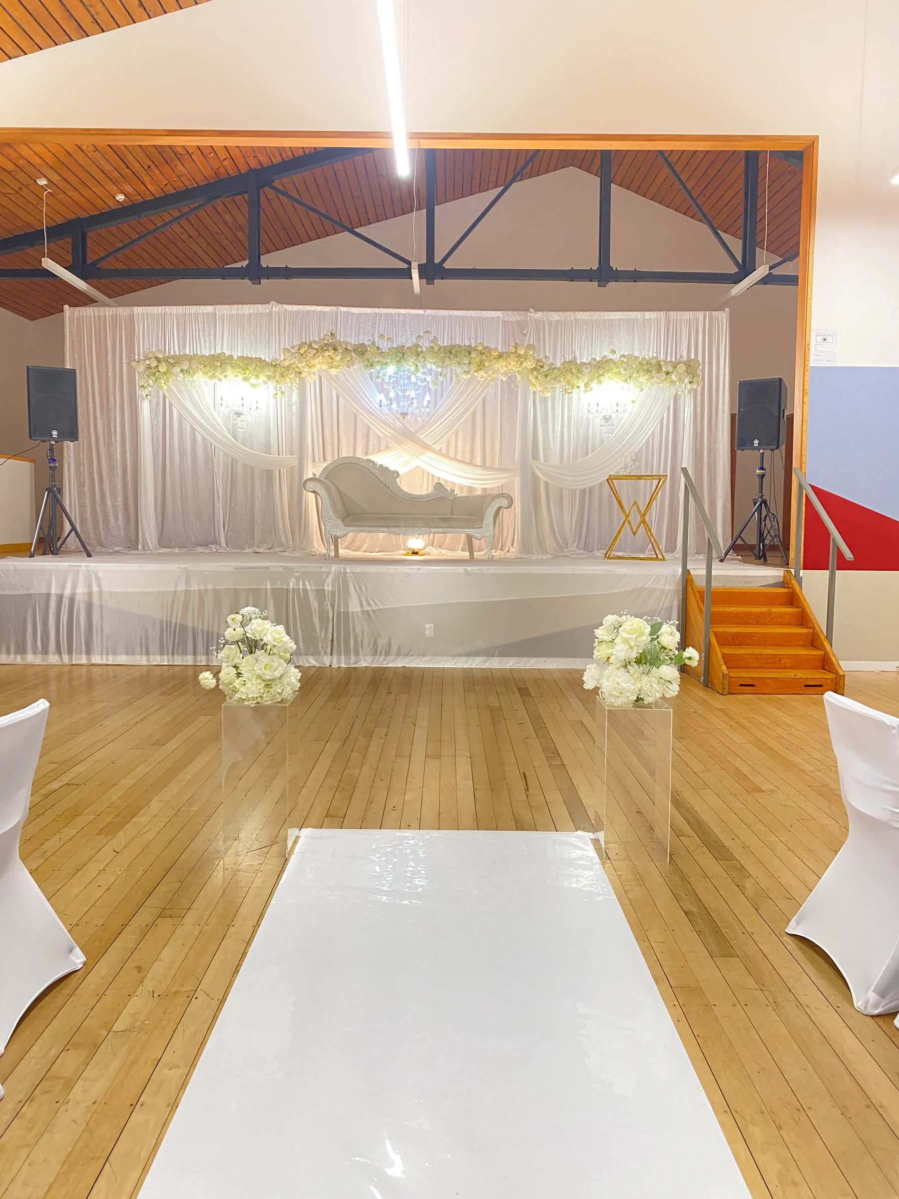 Ceremony styling by Decoration X: Mirror aisle between acrylic plinths of white roses to a white chaise under crossed chiffon swags and white floral garlands, beneath an exposed timber roof.