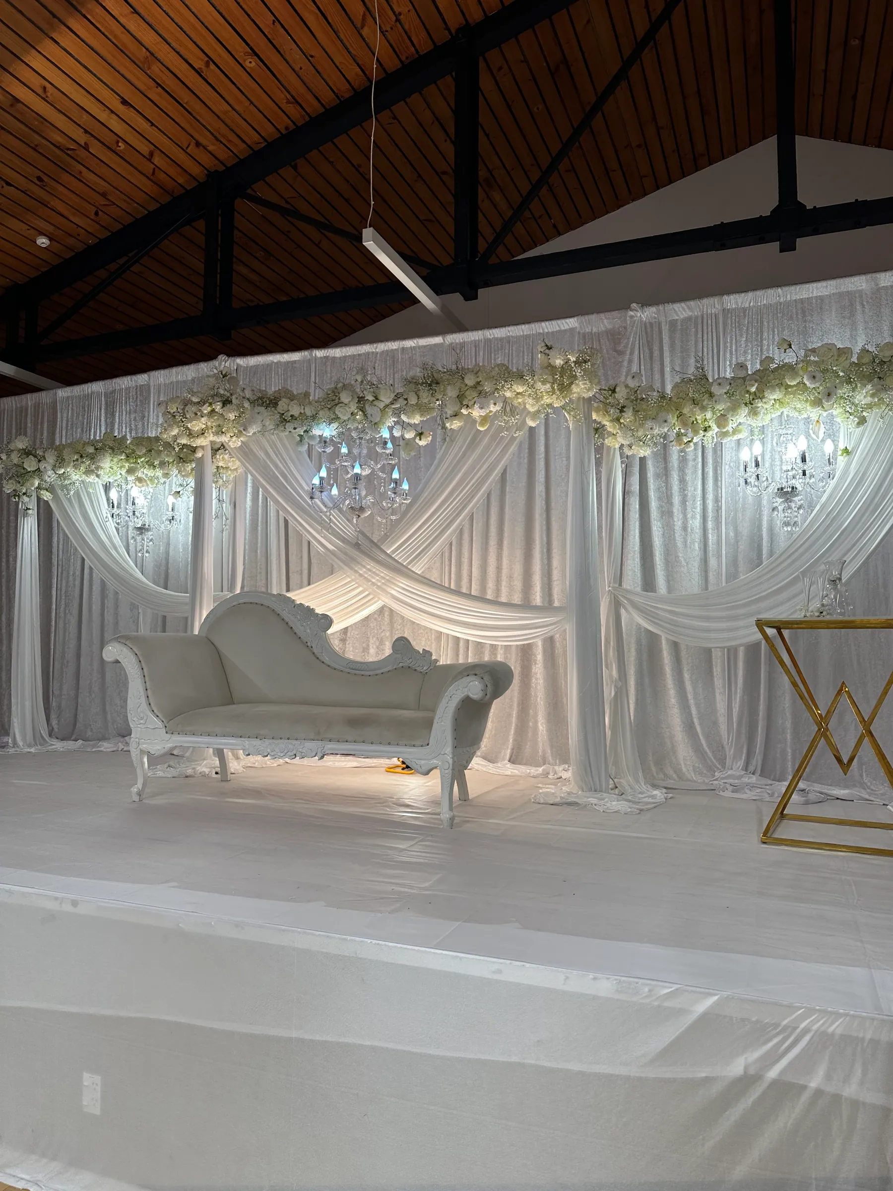 Stage styling by Decoration X: White chaise beneath crossed white chiffon swags and garlands of white roses and hydrangea, crystal chandeliers and a gold geometric stand beside it.