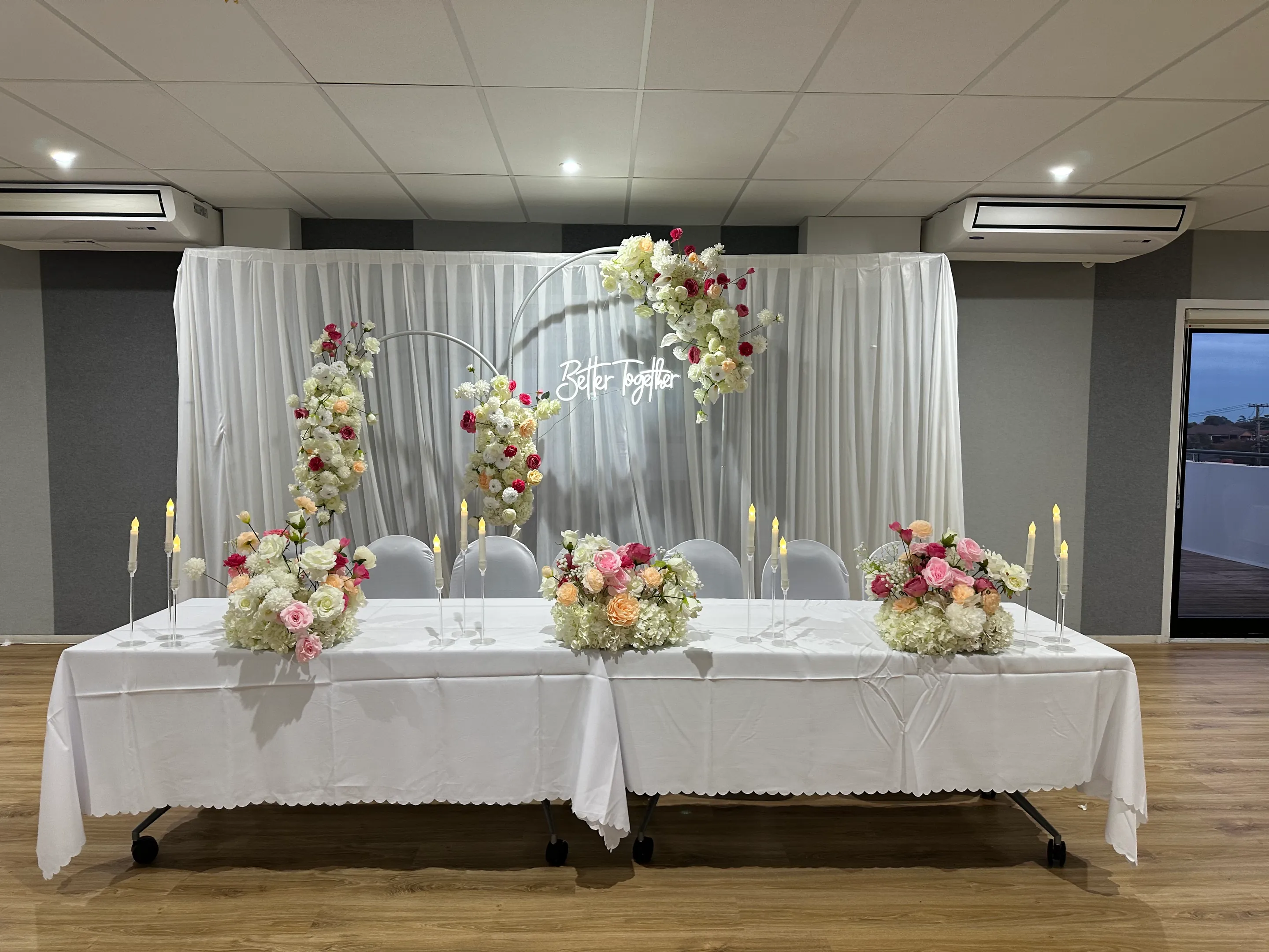 Stage styling by Decoration X: White-clothed head table with pink, peach and white floral mounds and taper candles before white hoops of roses and a Better Together neon on sheer drapes.