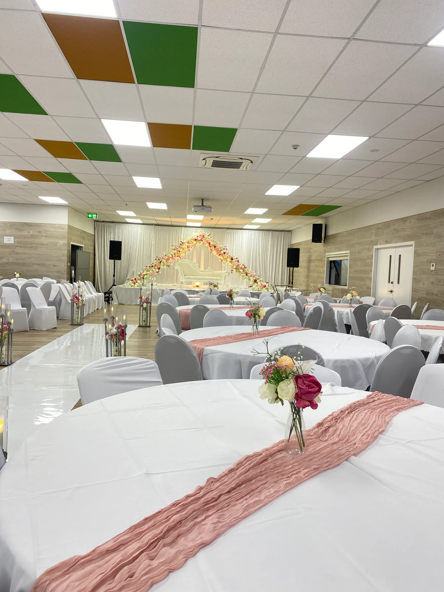 Reception styling by Decoration X: Round tables in white cloths with dusty-pink cheesecloth runners and bud vases of roses, a mirror aisle of candle stands leading to a floral-arched sofa stage.