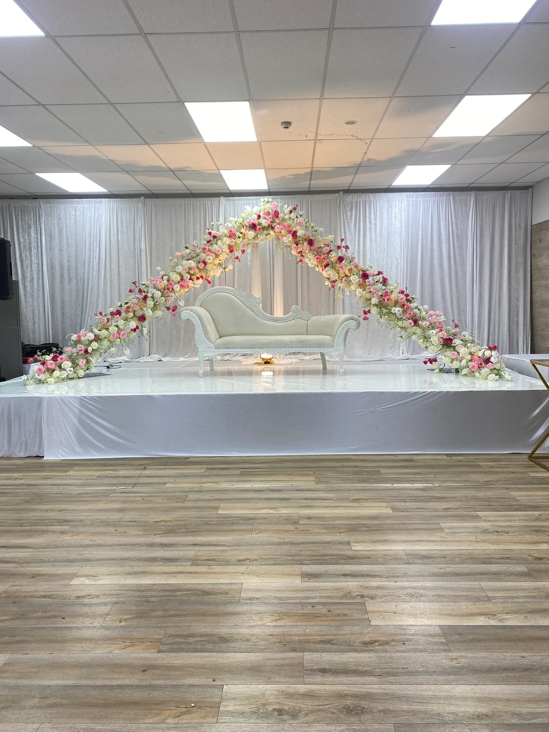 Stage styling by Decoration X: White chaise on a white-skirted stage beneath a wide triangular arc of pink, peach and white roses, white drapes behind and a small candle at its feet.