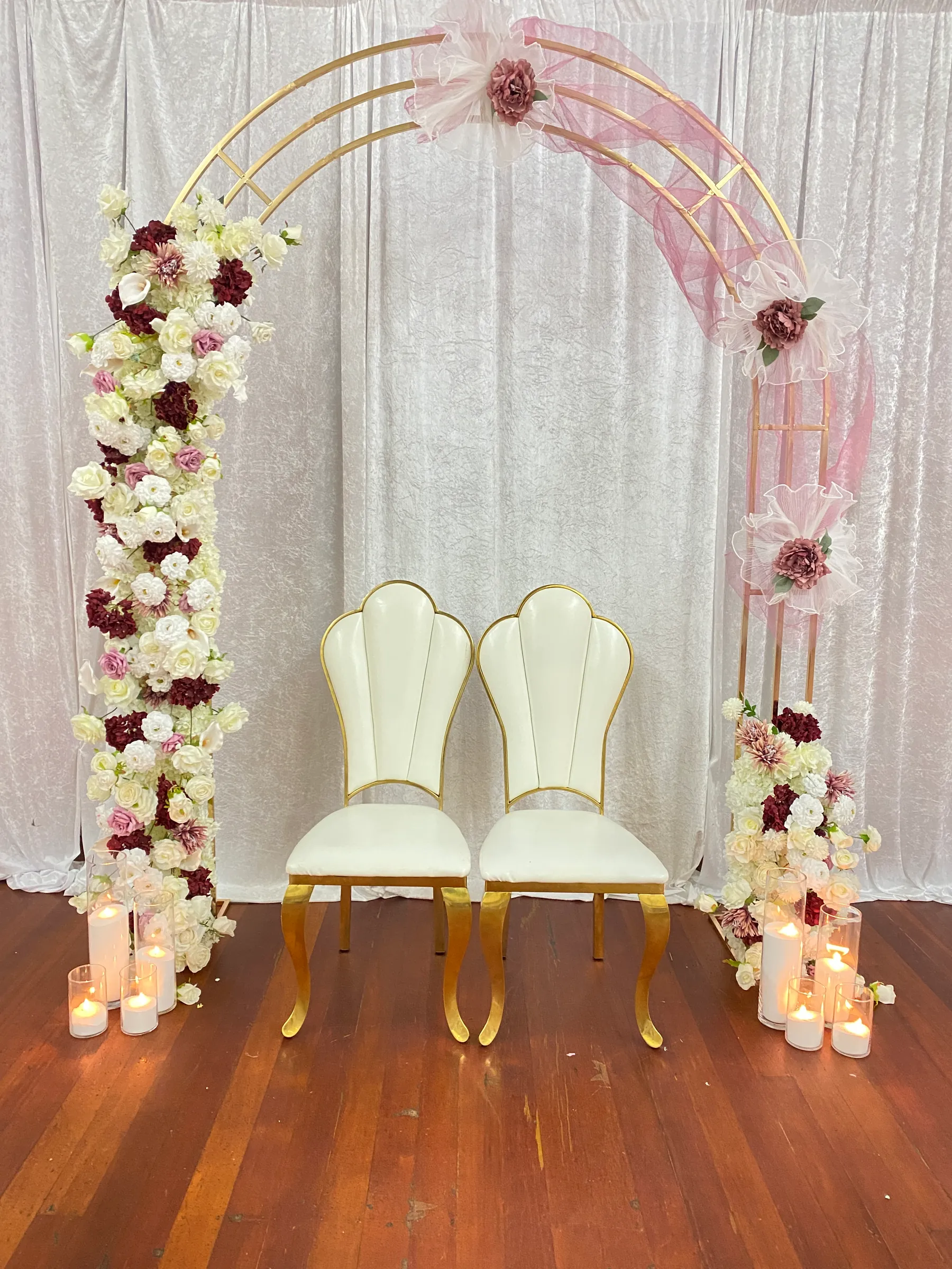 Stage styling by Decoration X: Gold triple arch climbing with ivory, burgundy and blush roses, a pink tulle swag with fabric blooms, two white shell-back chairs on gold legs, candles.