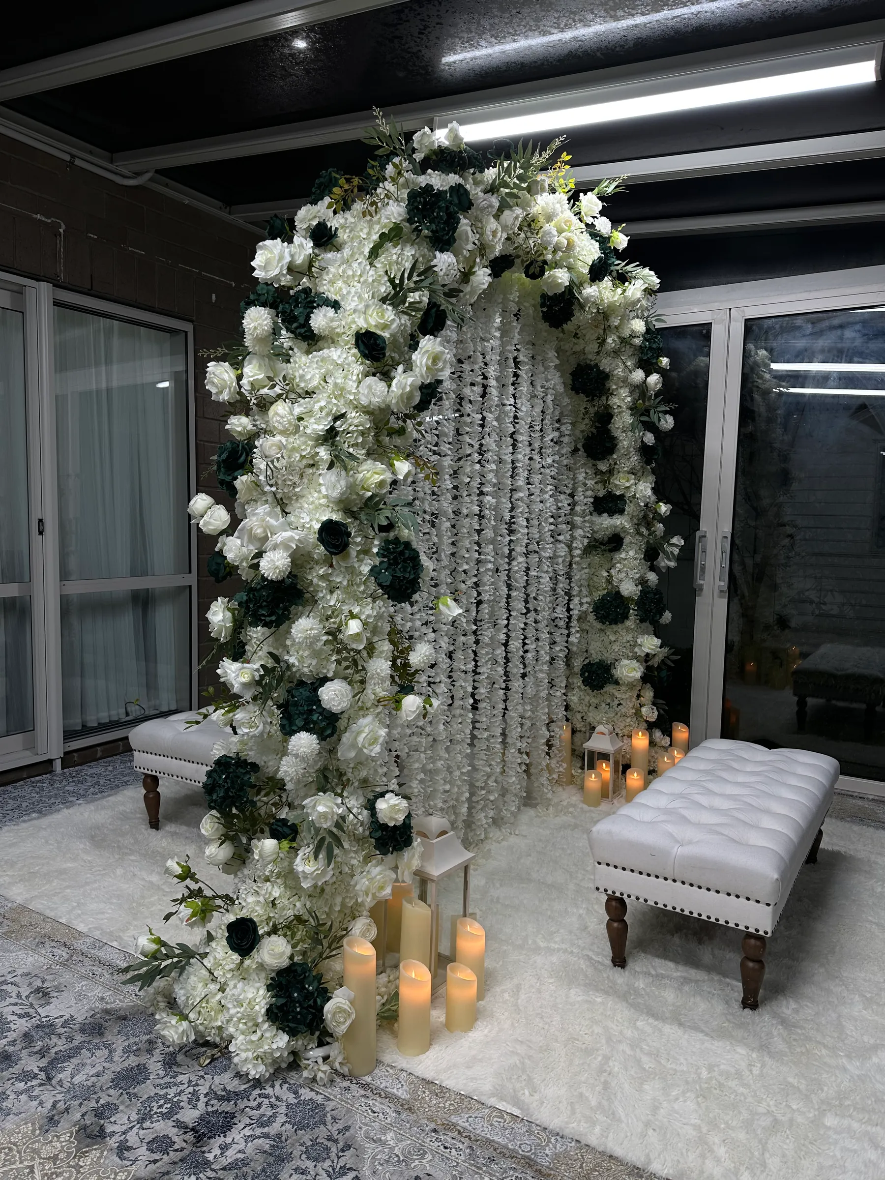 Stage styling by Decoration X: Arch of white and dark green roses around a white wisteria curtain, two tufted white benches on a shaggy white rug, lanterns and LED pillar candles.