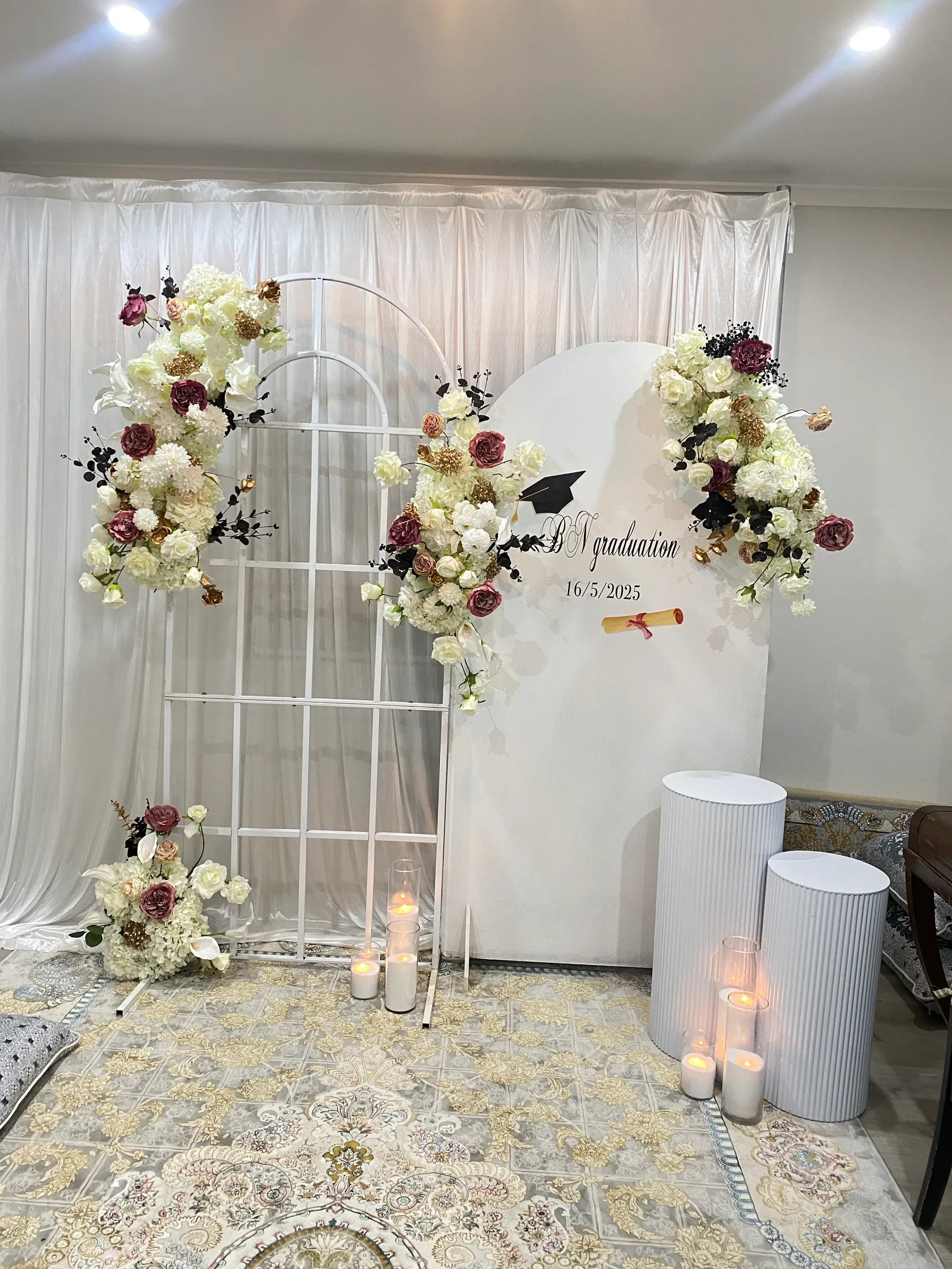 Graduation styling by Decoration X: White grid arch and arched panel with a mortarboard decal, ivory and burgundy roses with black and gold accents, fluted white plinths and candles.