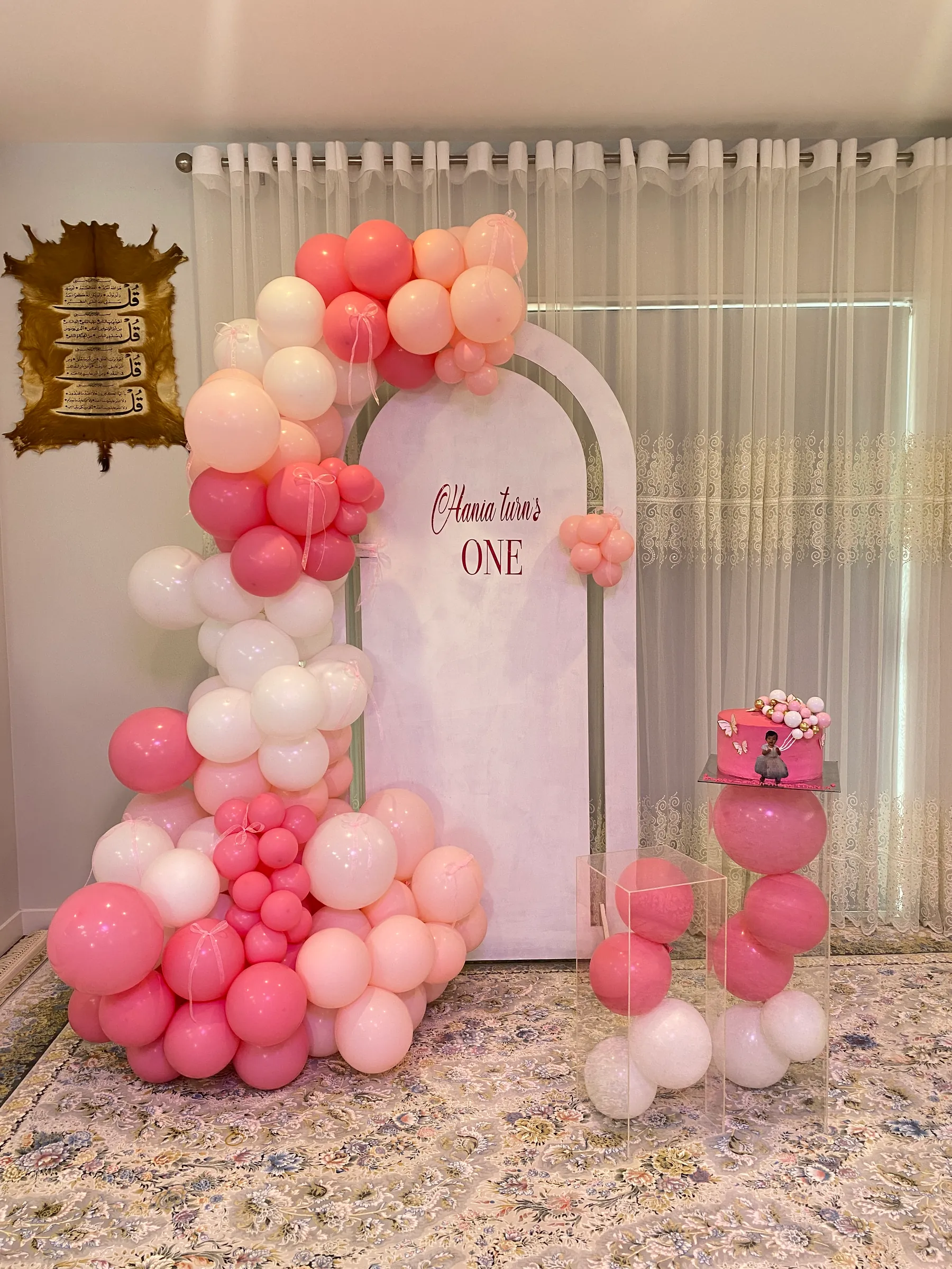 Birthday styling by Decoration X: Pink, peach and white balloon garland around a white arched panel with a first-birthday sign, acrylic boxes of balloons and a pink cake on a plinth.