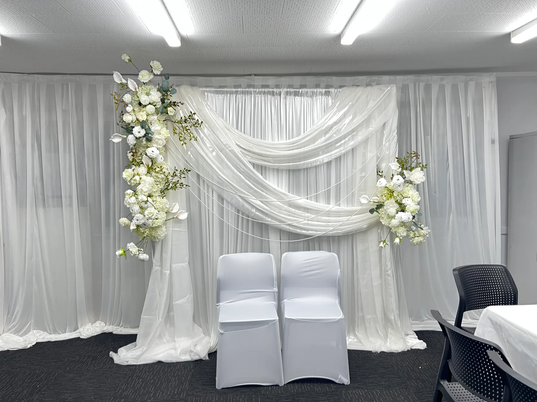 Stage styling by Decoration X: Two white-covered chairs before swagged white chiffon on sheer drapes, a cascade of white roses and hydrangea at one corner and a smaller spray opposite.