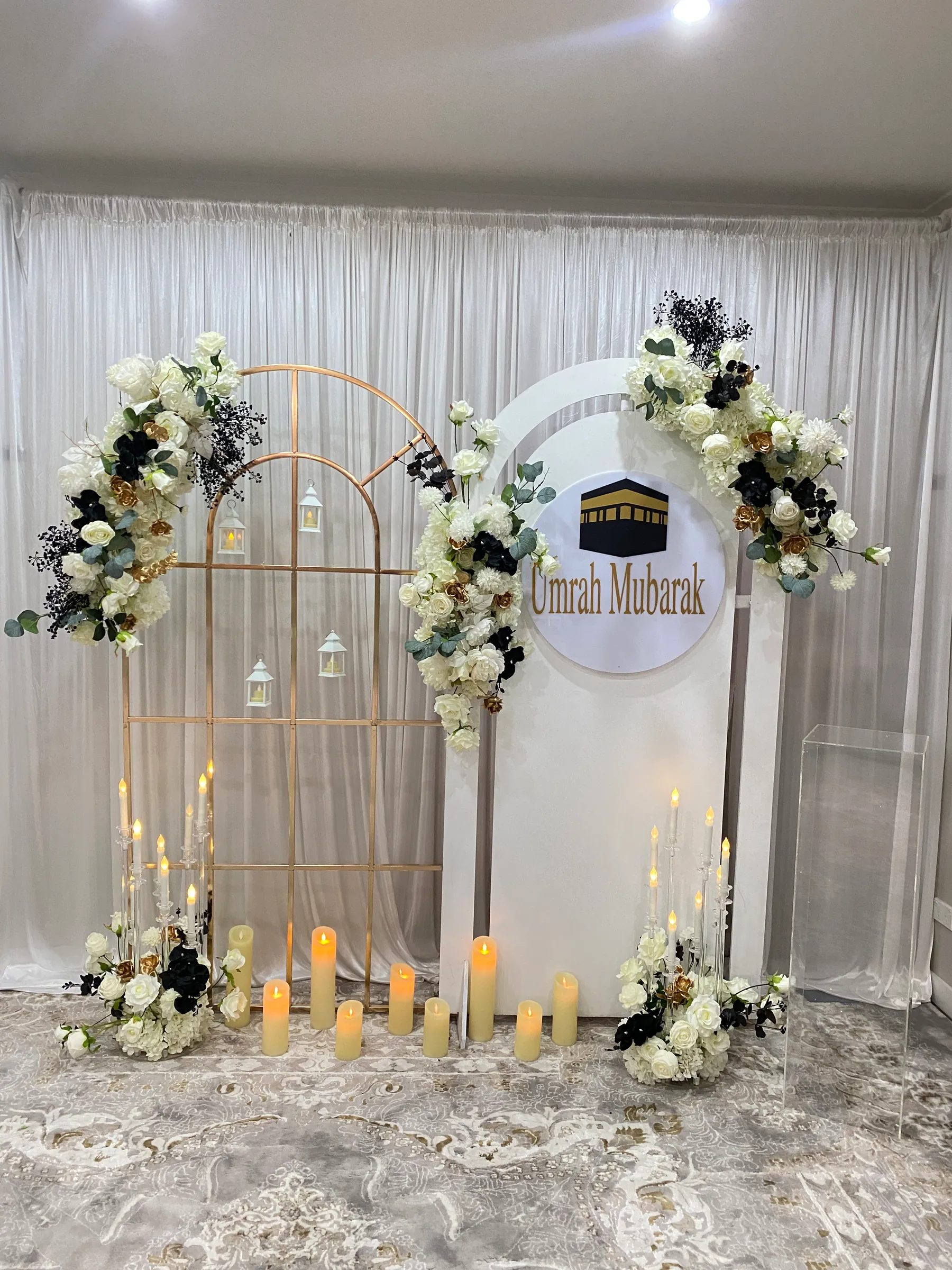Cultural styling by Decoration X: Gold grid arch hung with lanterns beside a white arched panel and Umrah Mubarak sign, white roses with black and gold accents, LED pillar candles.