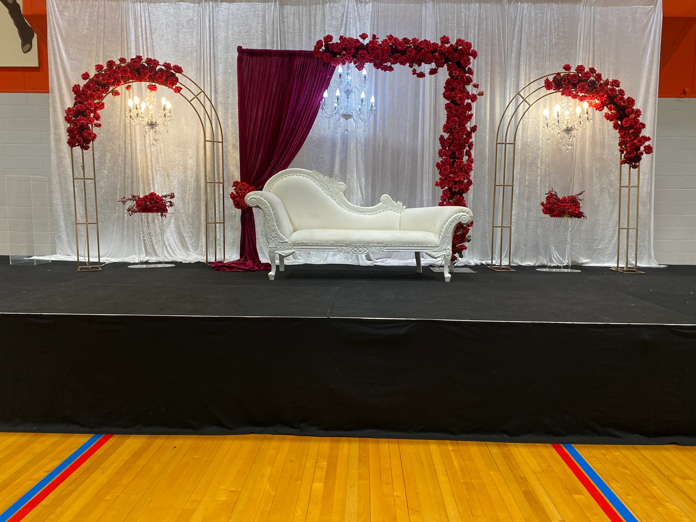 Stage styling by Decoration X: White chaise between gold arches heavy with red roses, a burgundy velvet drape, crystal chandeliers and acrylic plinths of red roses on a black stage.