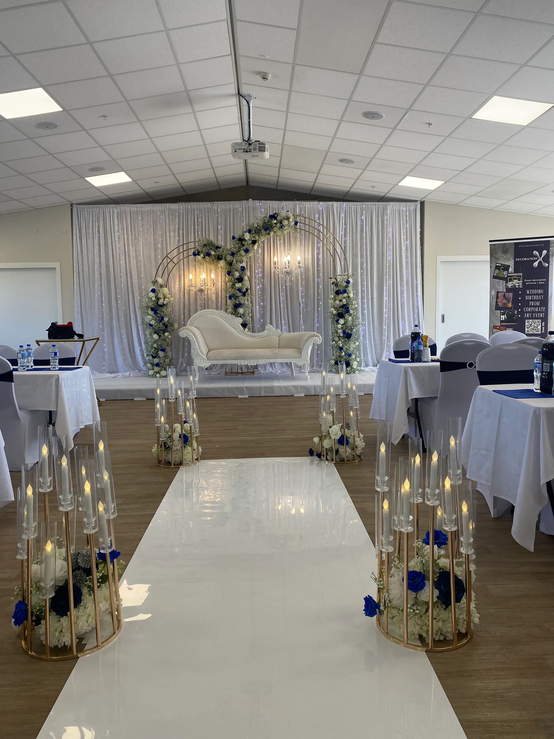 Ceremony styling by Decoration X: Mirror aisle between gold cage stands of LED candles and blue roses, leading to a white chaise under a gold double arch on a fairy-lit white drape.
