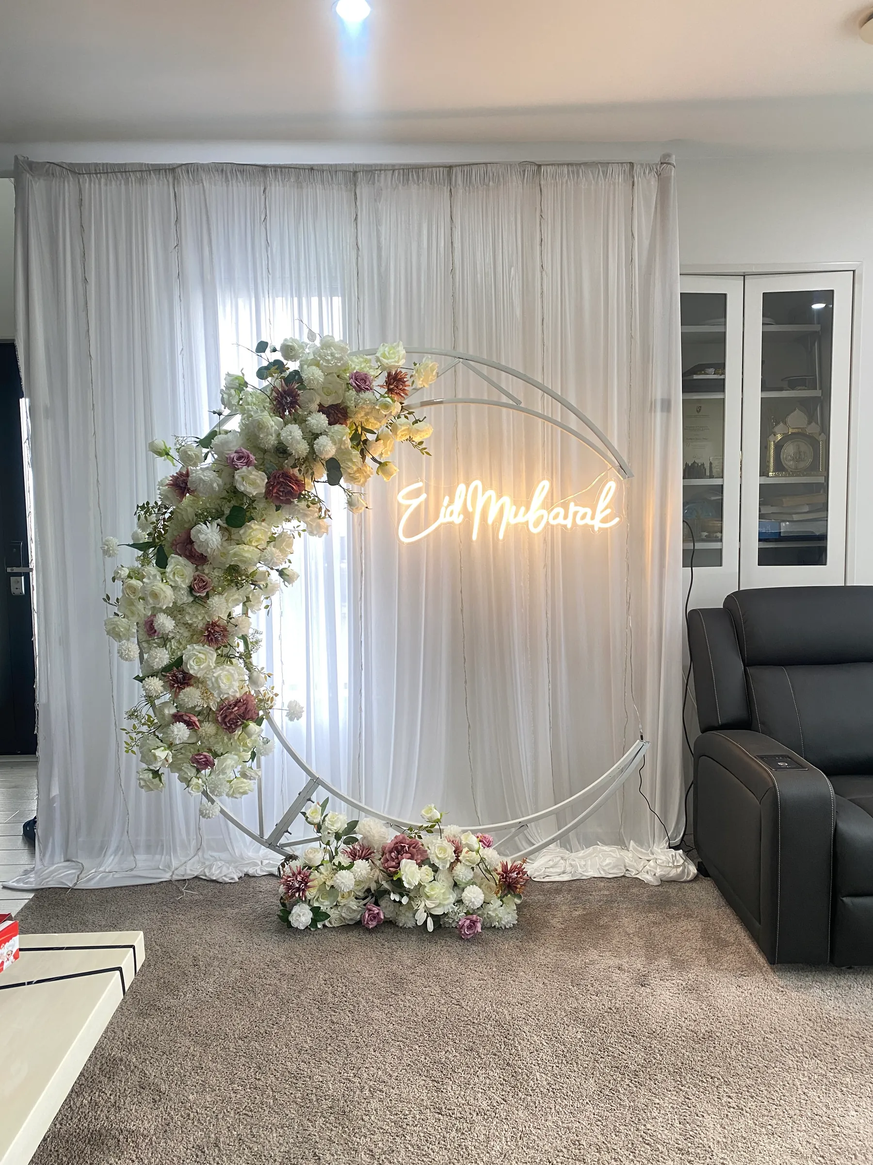 Cultural styling by Decoration X: White crescent-moon frame trailing ivory and dusky-pink roses before sheer white drapes, a neon Eid Mubarak sign glowing inside the curve.