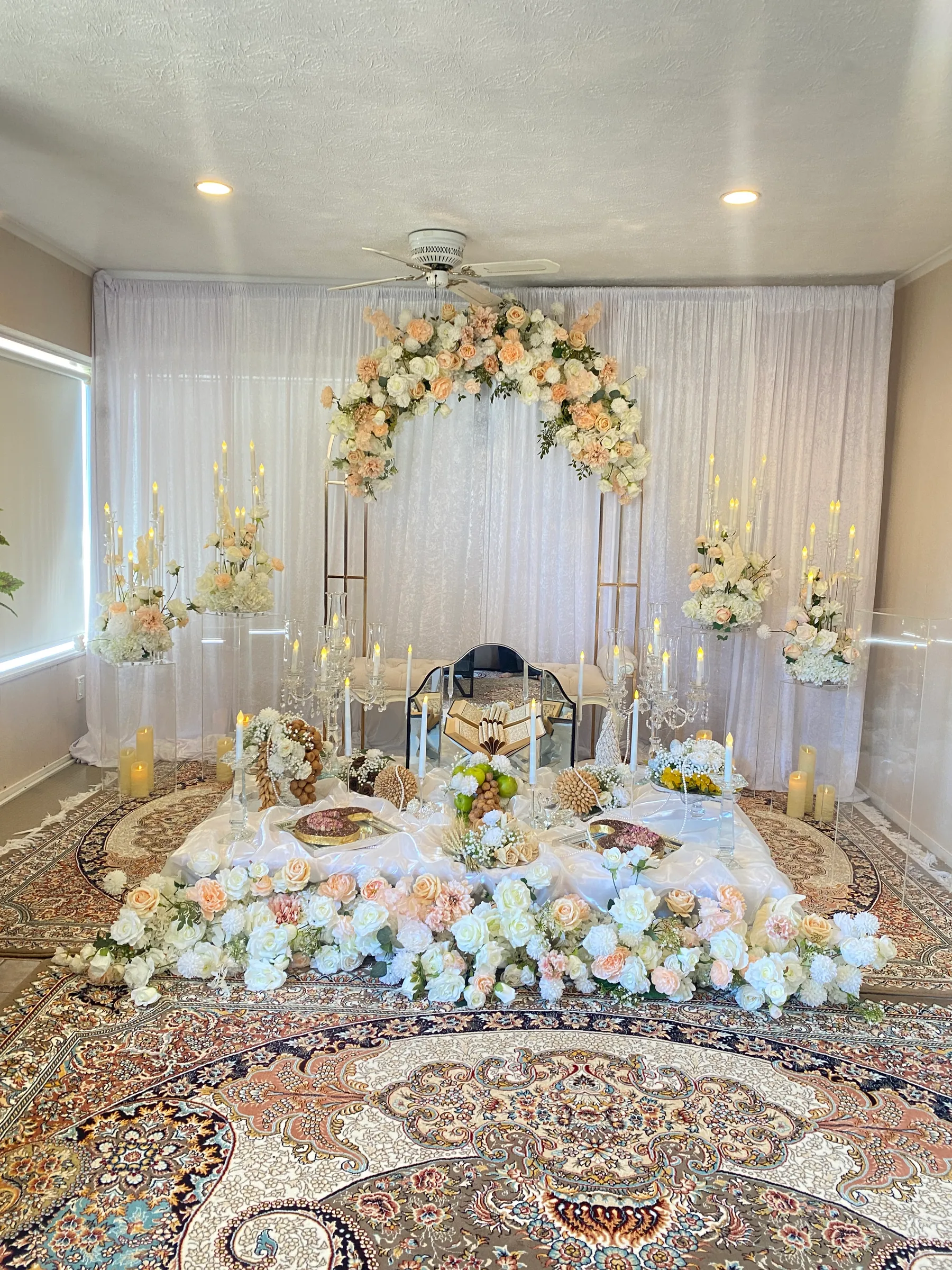 Cultural styling by Decoration X: Sofreh aghd on Persian rugs beneath a peach and white rose arch, crystal candelabras, a mirror, decorated breads and fruit, a rose garland along the front.