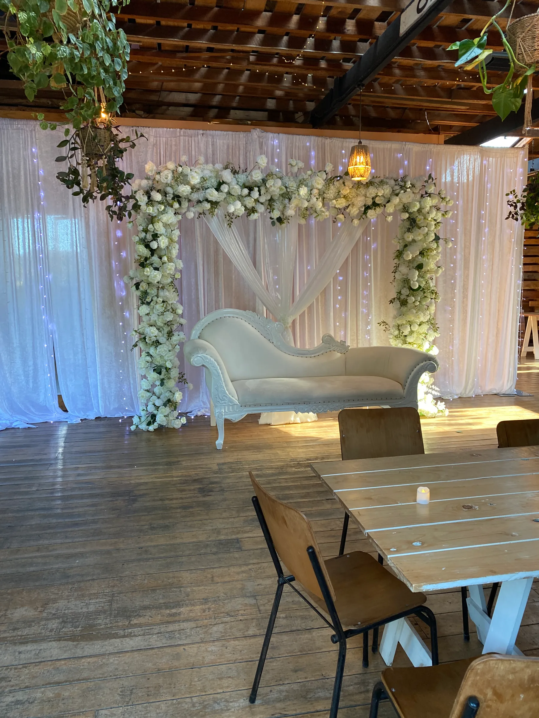 Stage styling by Decoration X: White rose square arch over crossed sheer drapes and an ivory chaise, fairy-lit white drapes behind, in a timber-floored venue with hanging plants.