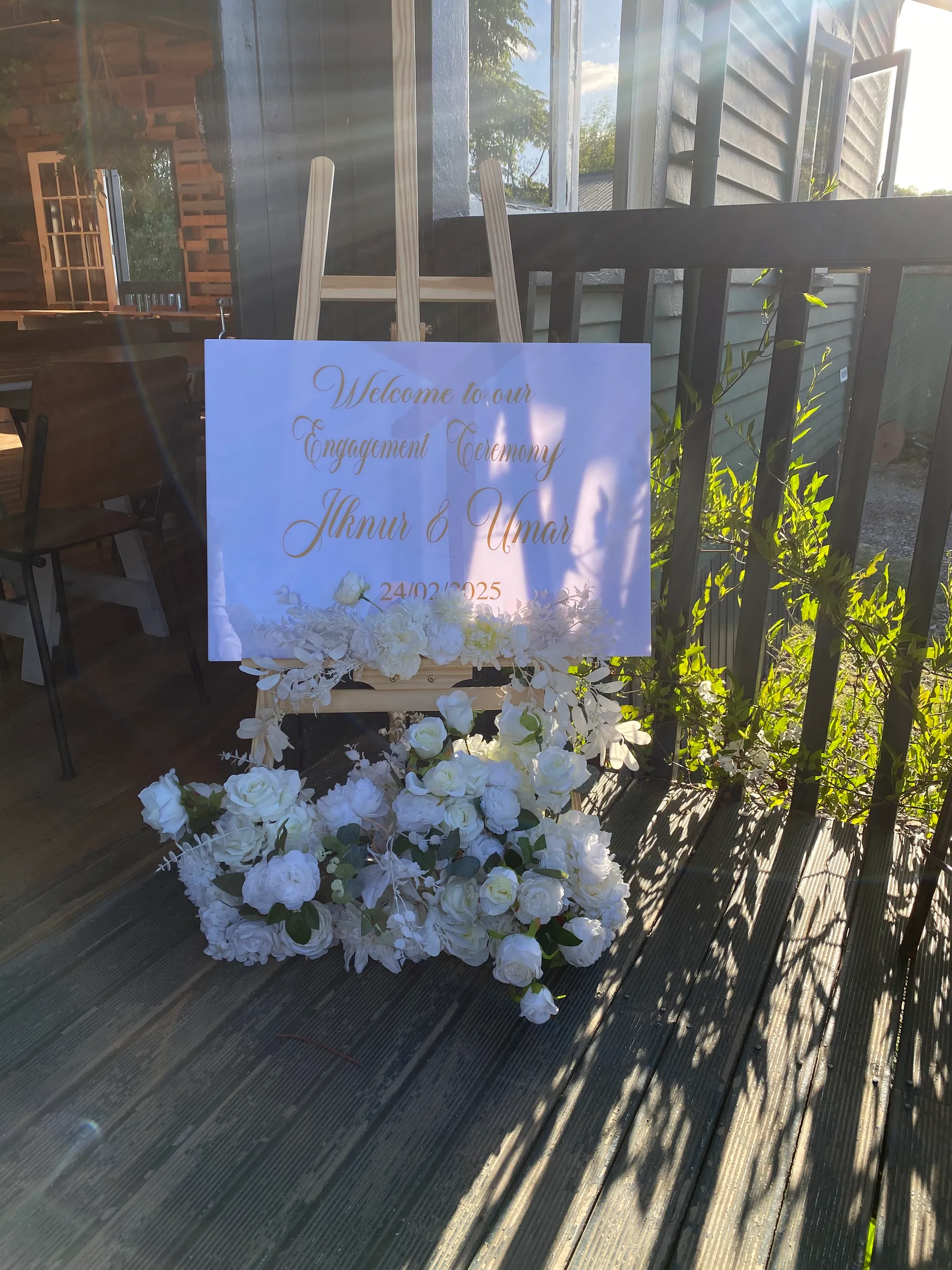 Engagement styling by Decoration X: White acrylic welcome sign in gold script on a timber easel, white roses heaped at its foot, on a sunlit deck.