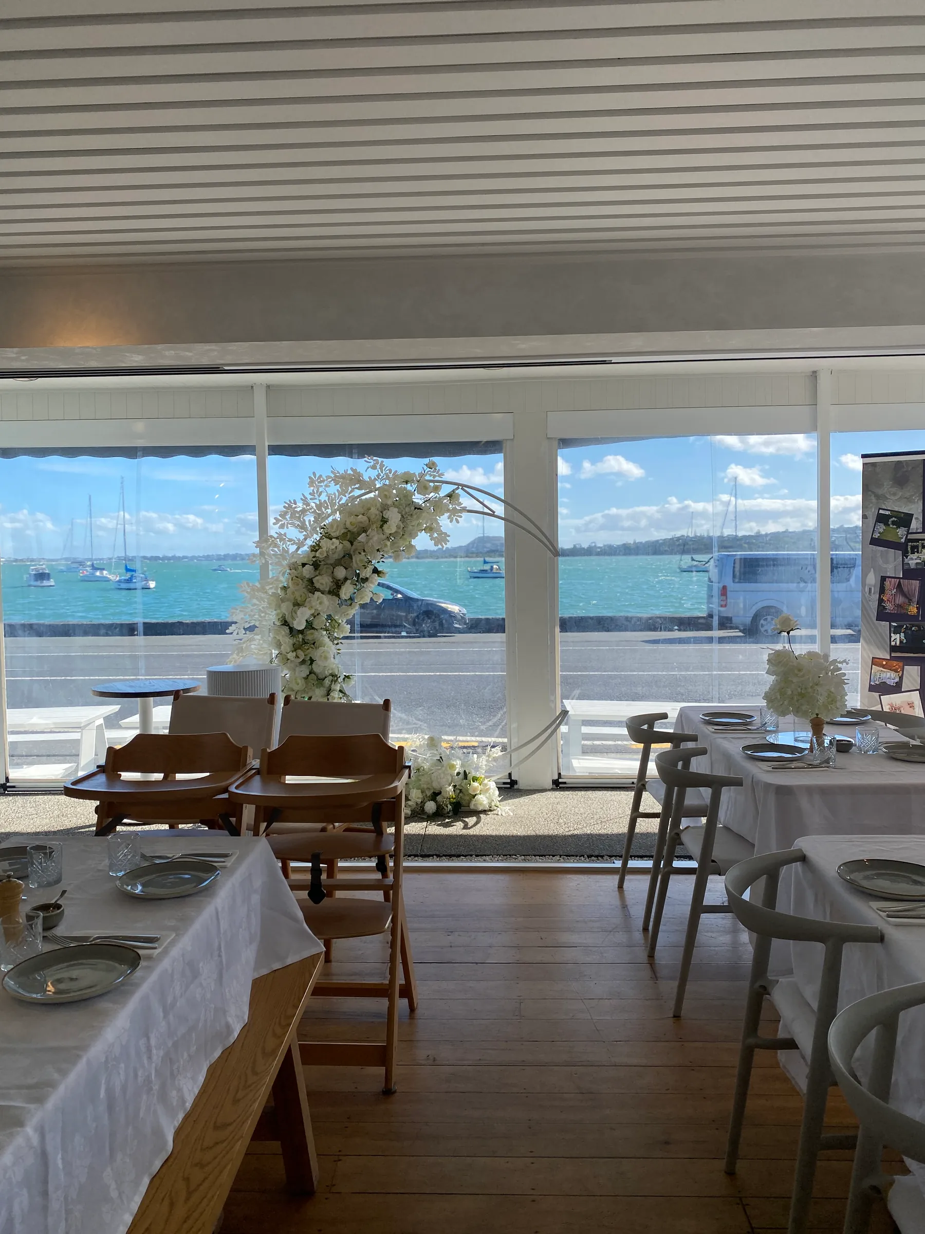 Reception styling by Decoration X: White floral half-moon hoop in a waterfront restaurant window overlooking moored yachts, tables set with white linen and grey plates.
